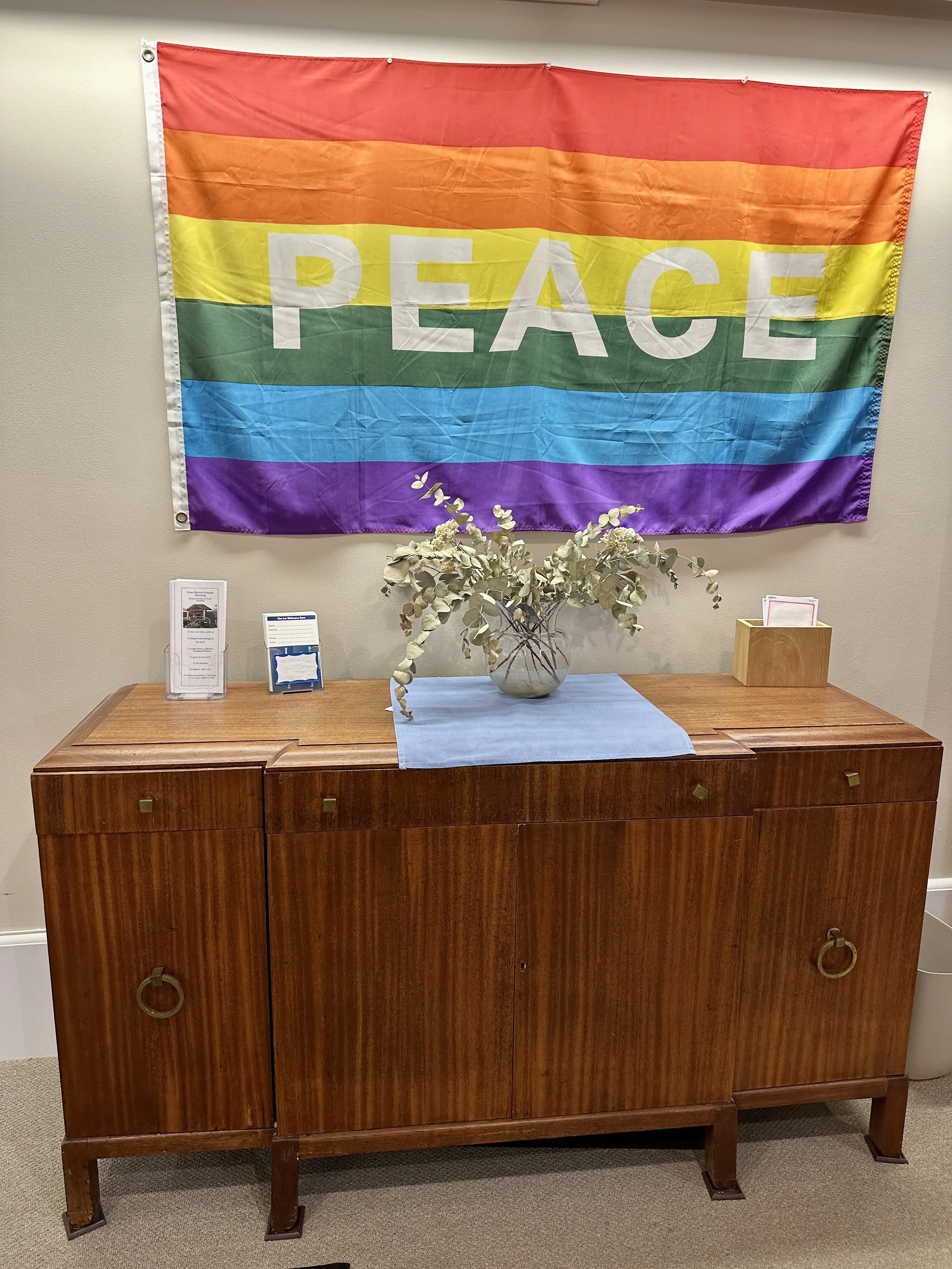 A rainbow flag with the word 'PEACE' hangs on a wall above a wooden sideboard with a vase of white flowers and informational pamphlets.