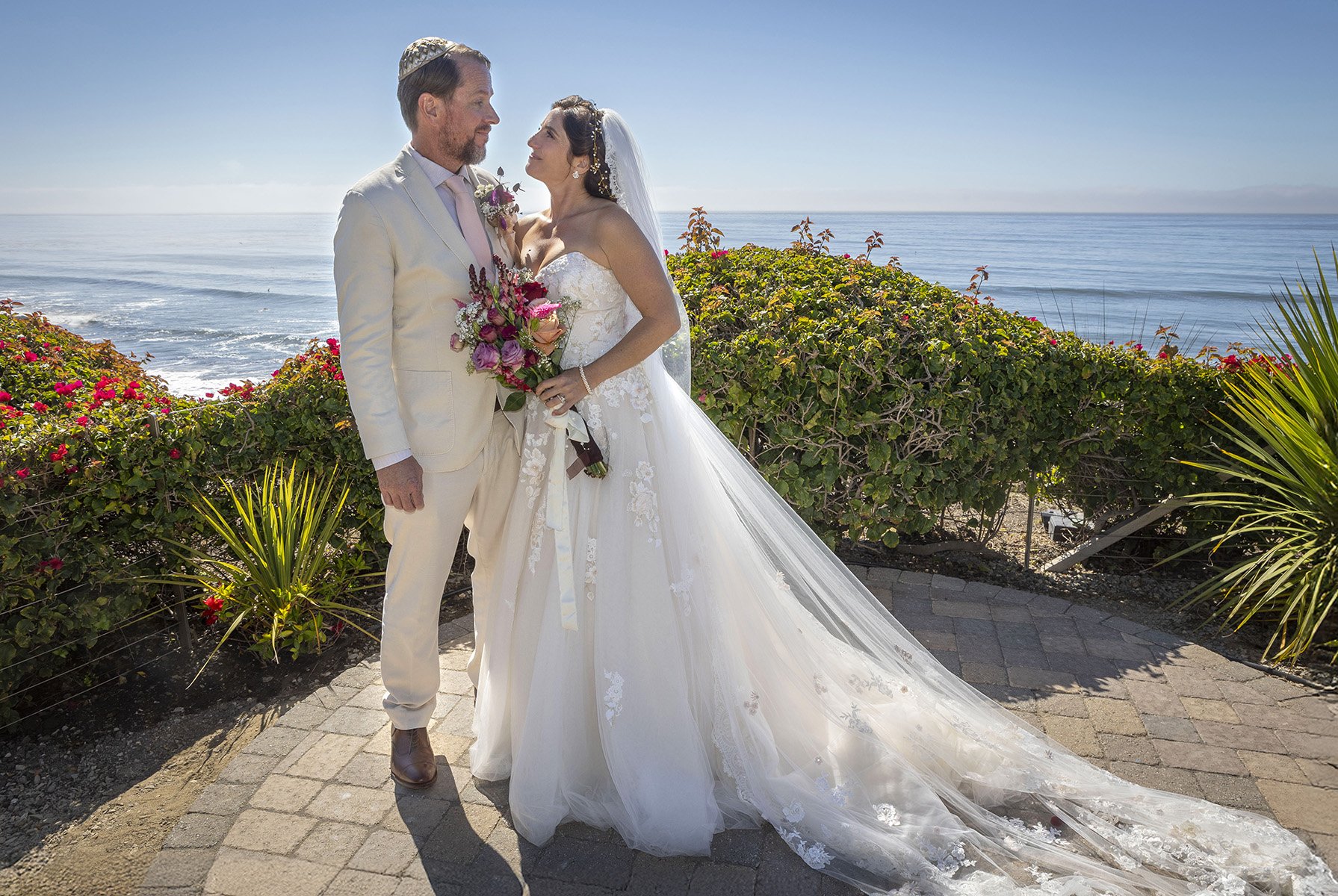 A bride and groom standing outdoors near the ocean, dressed in wedding attire, with the bride holding a bouquet of pink and purple flowers, and a lush green beachside background.