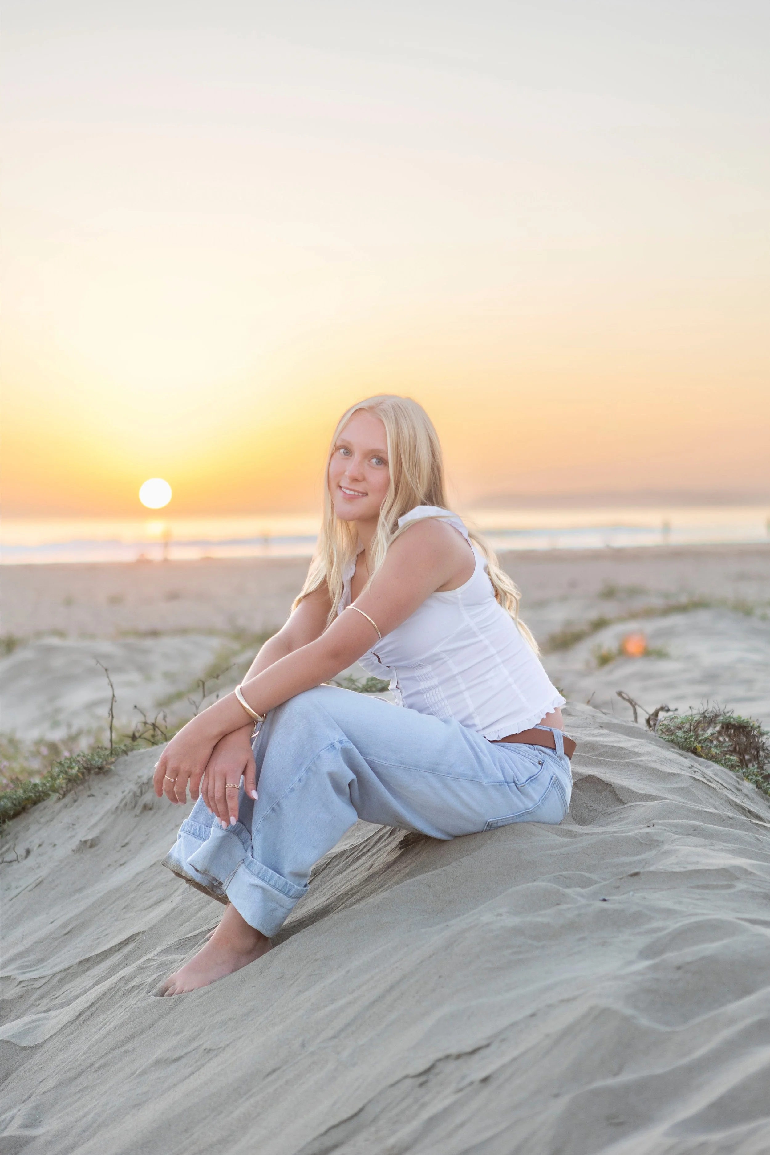 Young woman with long blonde hair sitting on a sandy beach during sunset, smiling and looking at the camera, wearing a white sleeveless top and light blue jeans.