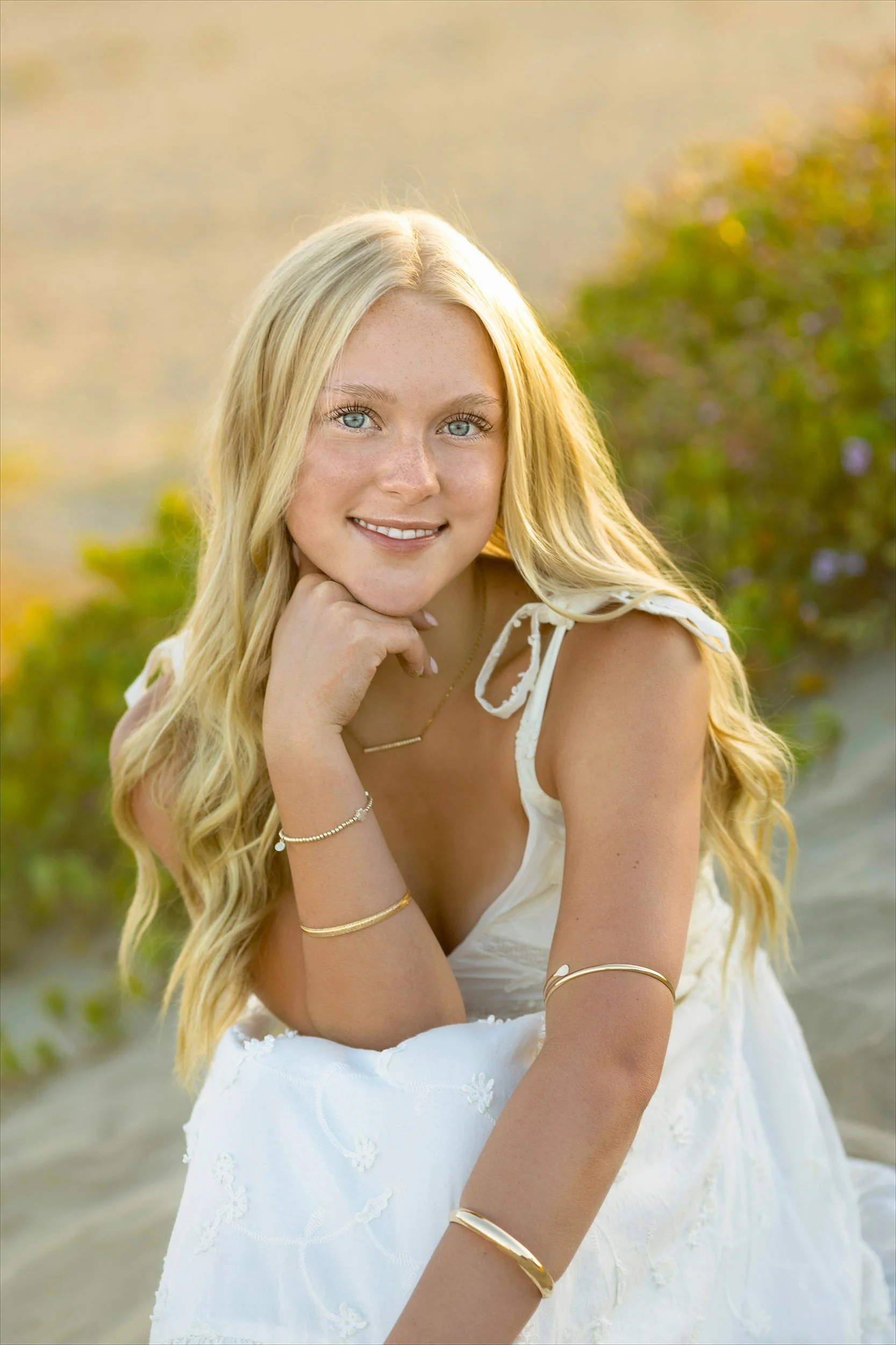 A young woman with long blonde hair and blue eyes smiling outdoors in a white dress, accessorized with gold jewelry, with greenery and a blurred background.