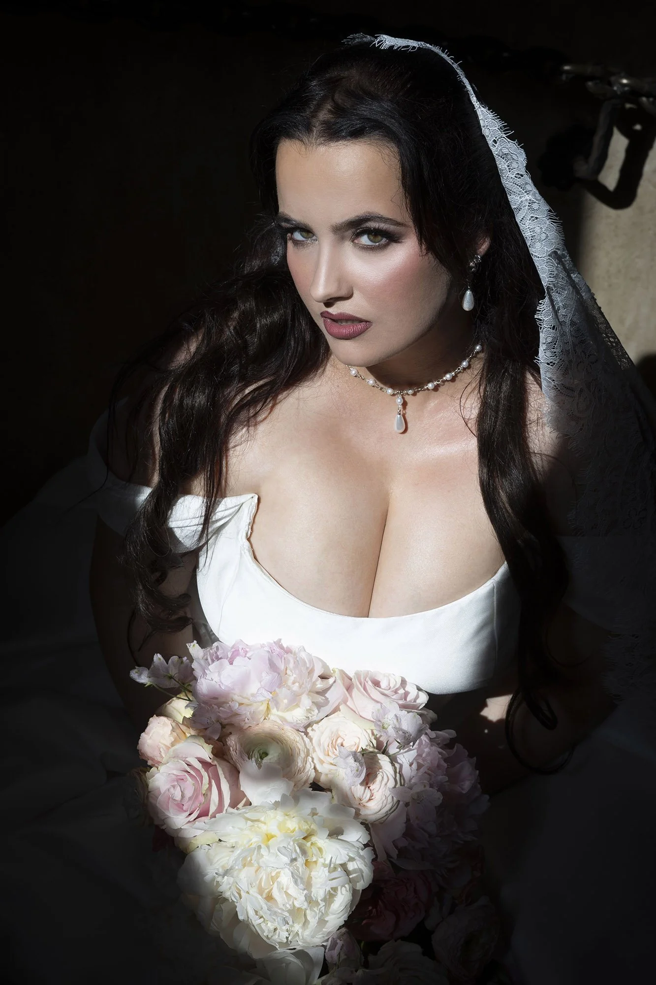 A woman in a bridal dress with a veil, holding a bouquet of peonies and roses, looking directly at the camera with a serious expression.