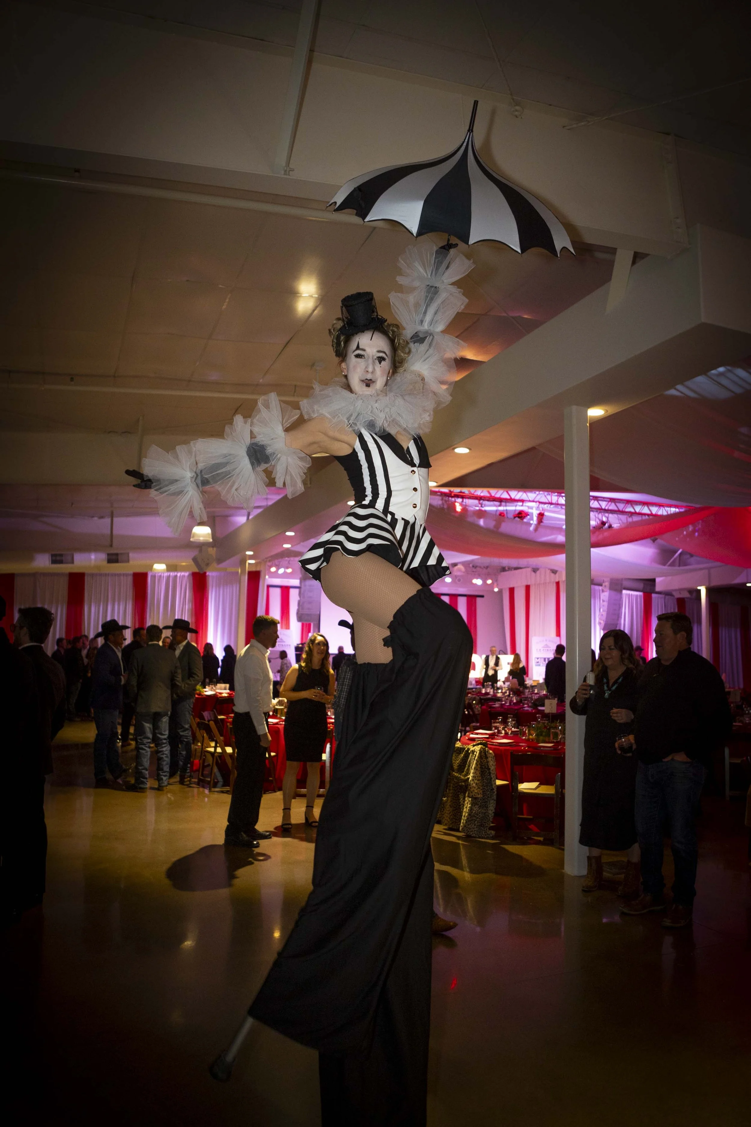 A woman dressed as a mime in a black and white striped outfit, balancing on stilts at an indoor event with people socializing in the background. She holding an umbrella and is surrounded by pink-themed decorations.