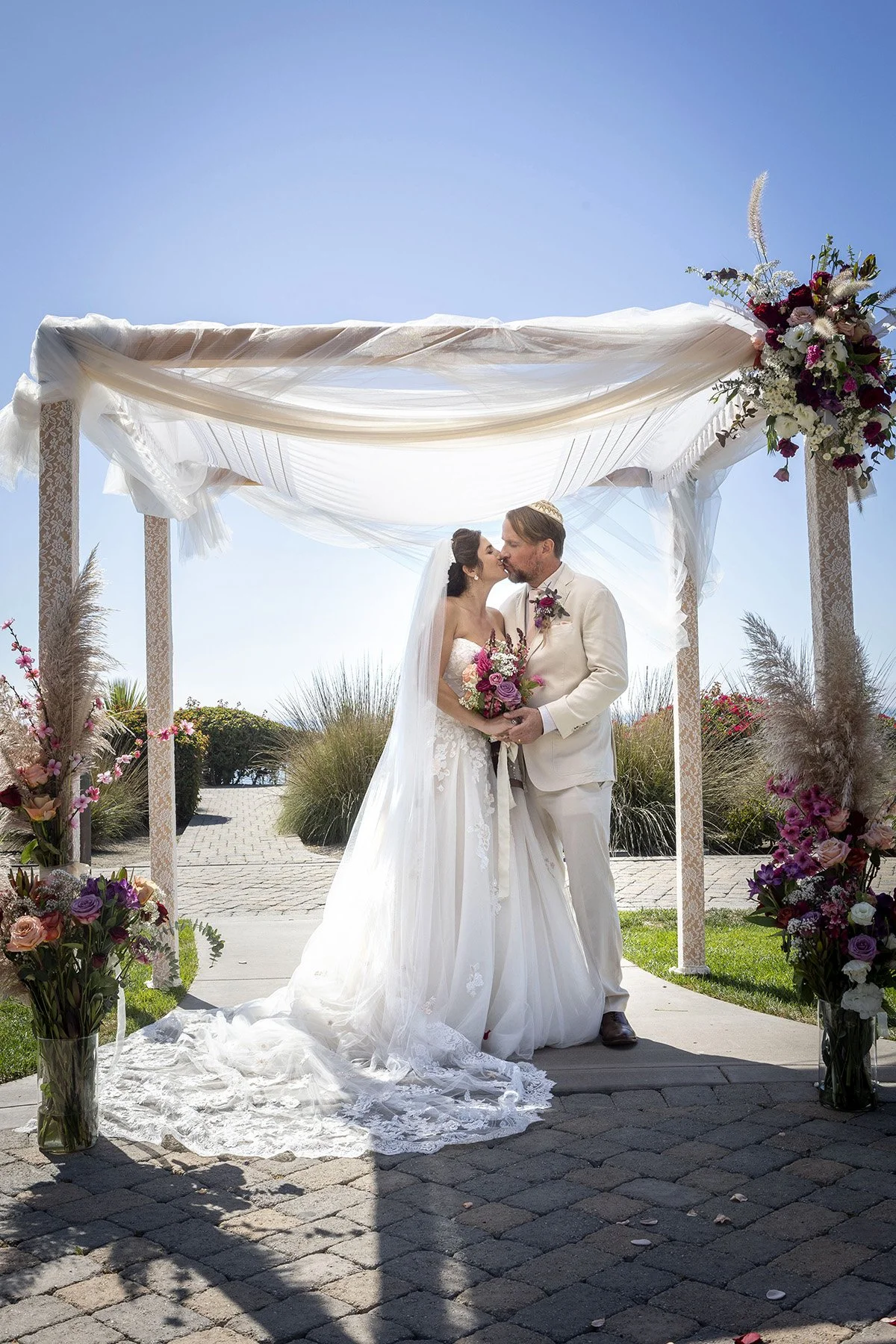 A bride and groom kissing under a decorated wedding canopy on a sunny day, holding a bouquet of pink and purple flowers.