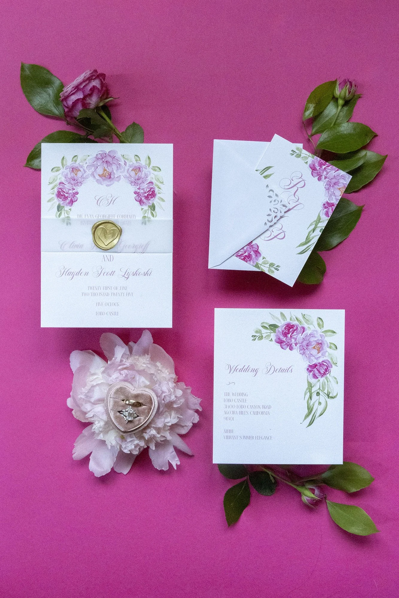 Wedding invitation suite including an invitation card with floral design and a wax seal, a folded RSVP card, a floral-themed wedding details card, pink flower with rings attached, and greenery on a pink background.
