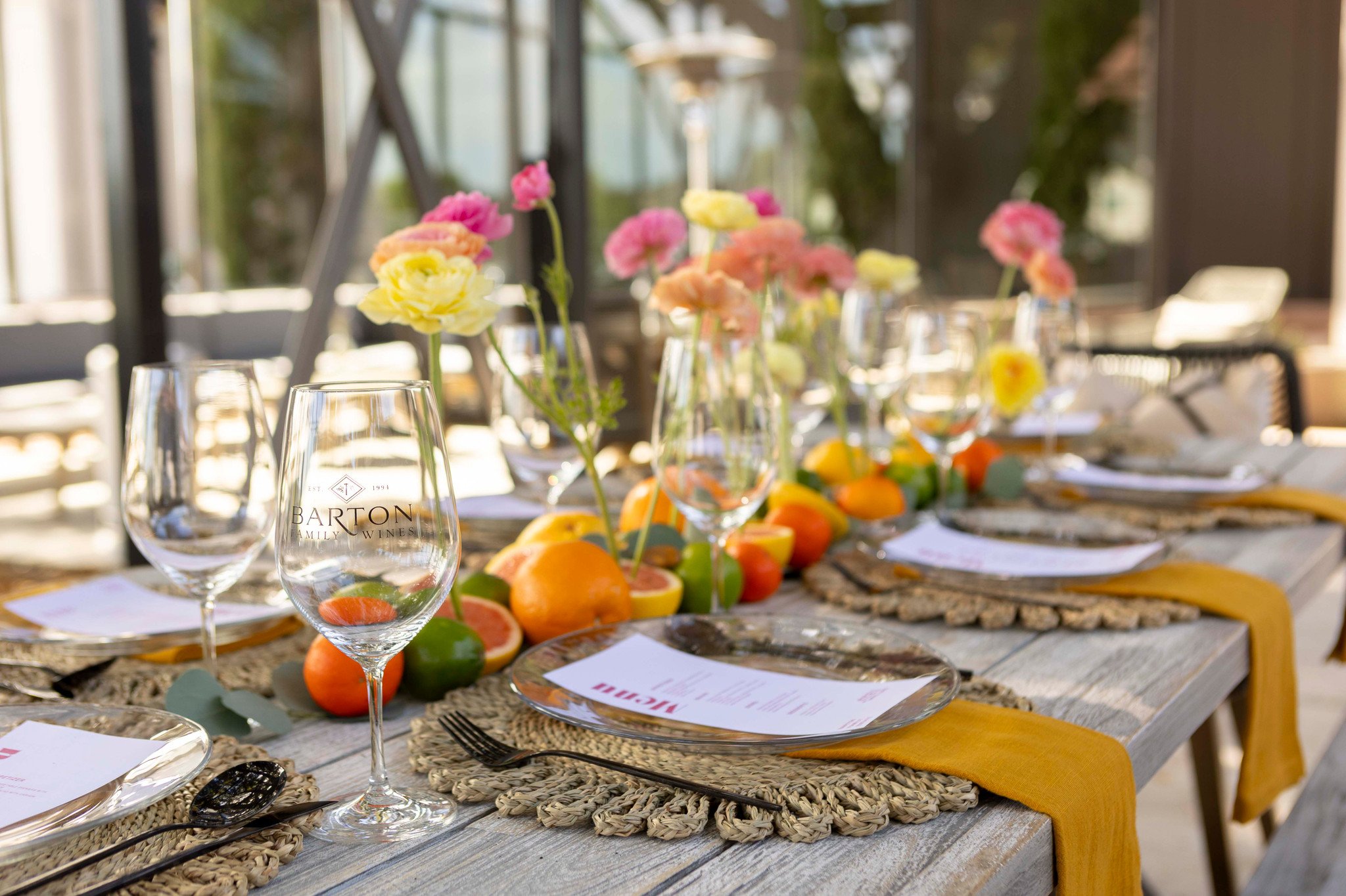 A well-decorated outdoor dining table with woven placemats, yellow napkins, glassware, colorful flowers, and an array of fresh citrus fruits in the center.