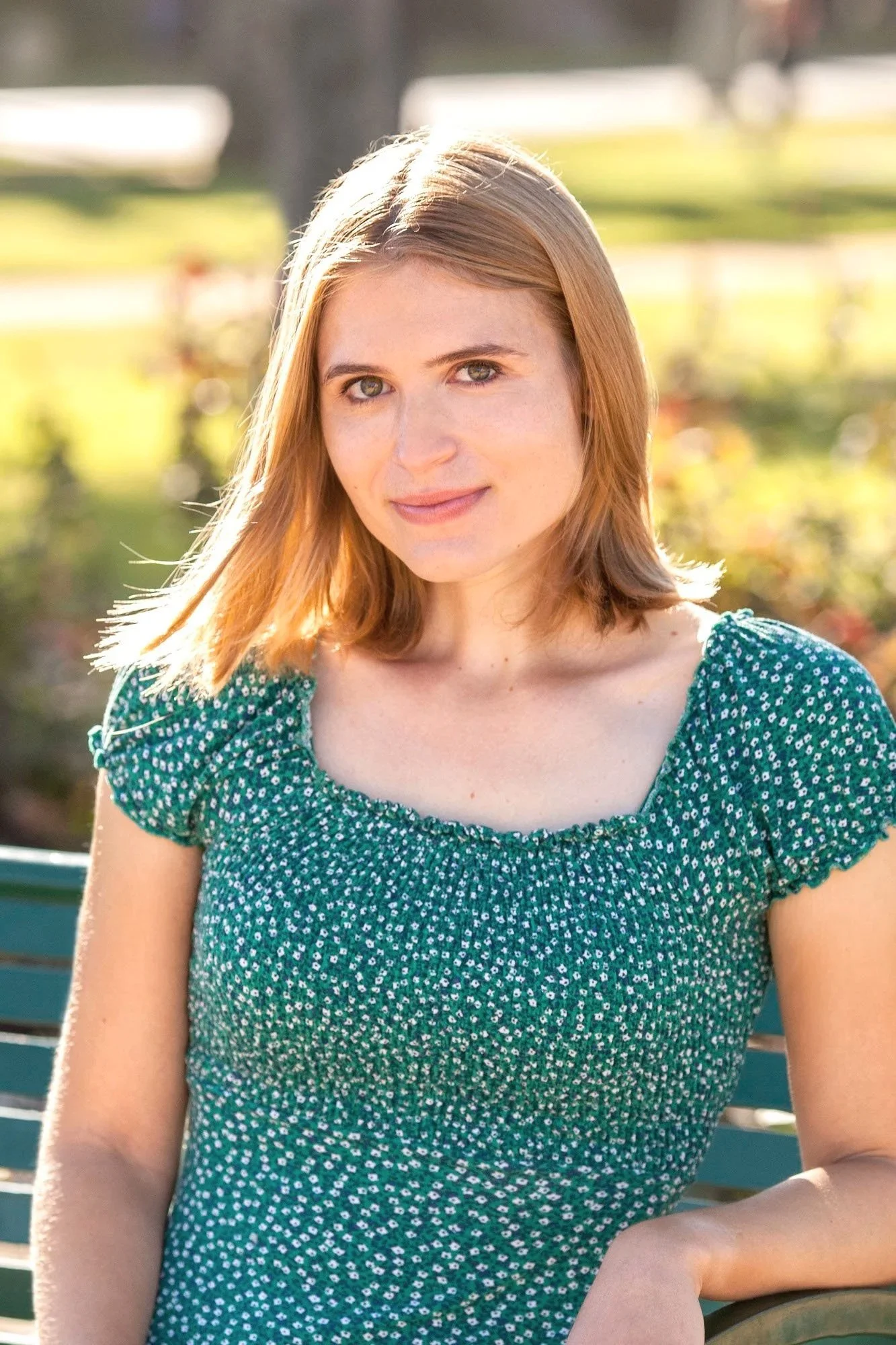 A woman with shoulder-length light brown hair sitting on a park bench, wearing a green patterned short-sleeve top, with a blurred green and yellow park background.