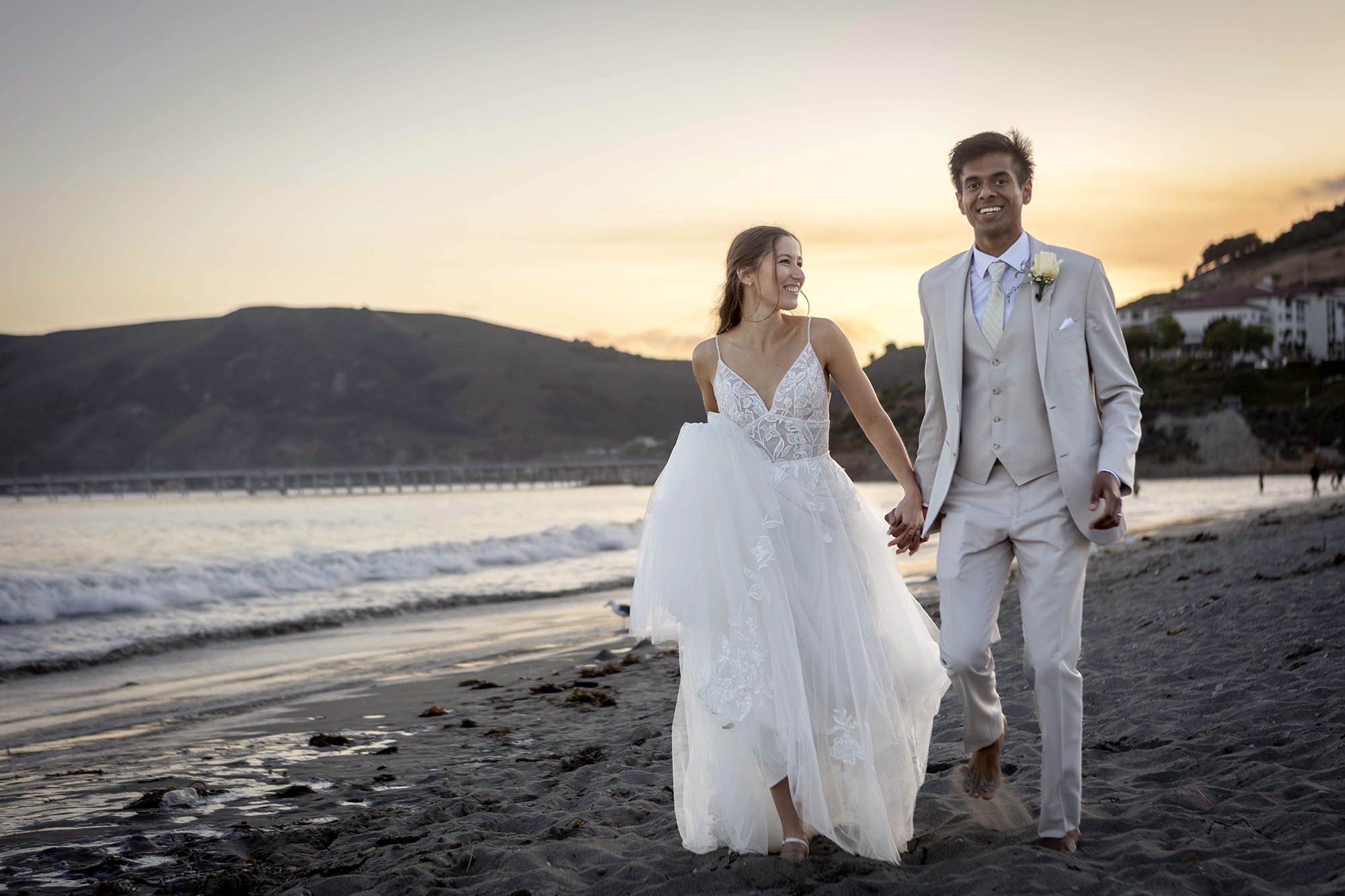 A newlywed couple walking barefoot on a beach during sunset, holding hands and smiling at each other. The bride is wearing a white wedding dress and the groom is in a white suit with a boutonniere.