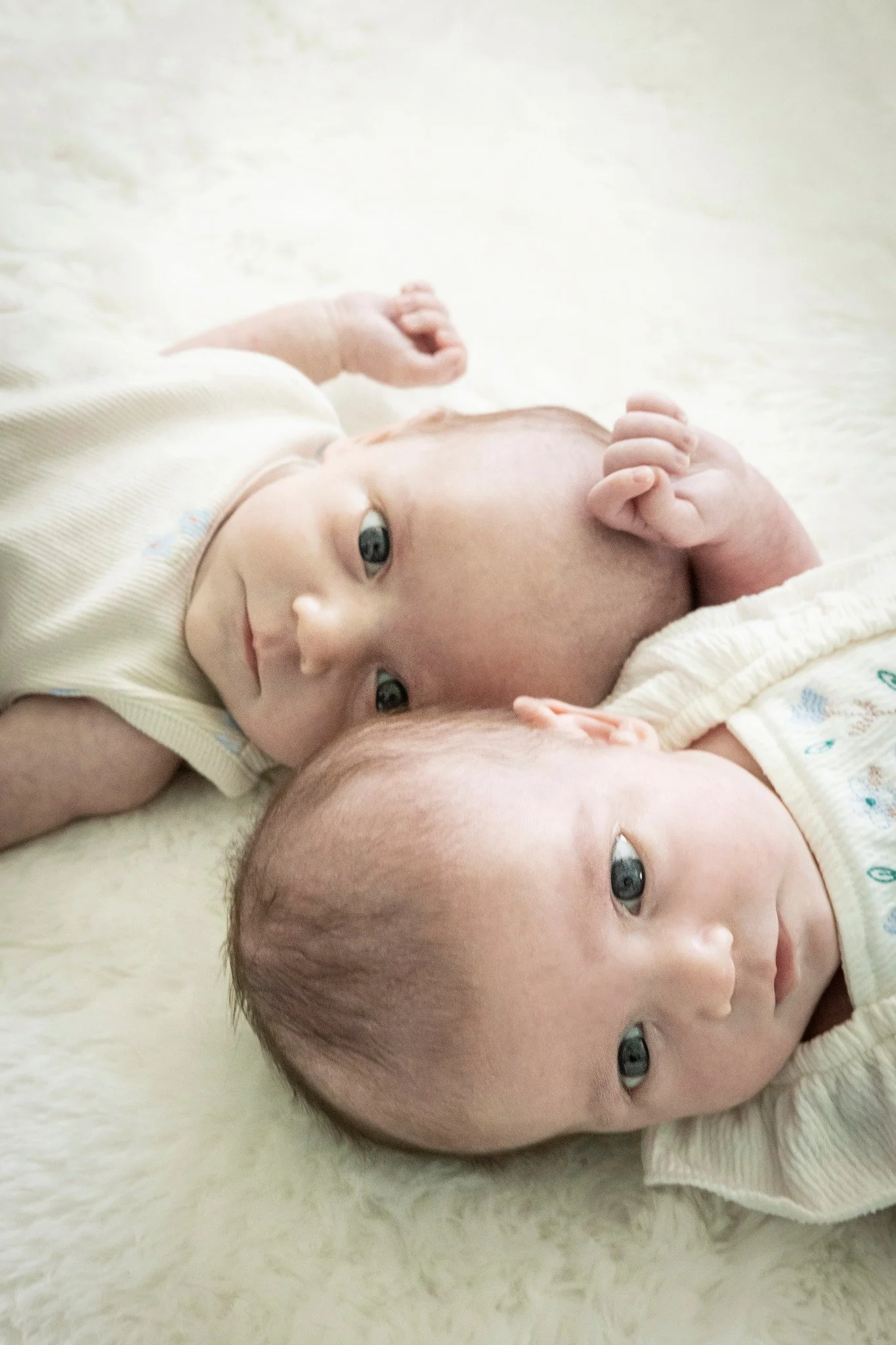 Close-up of two newborn babies lying on a soft, cream-colored surface. They have blue eyes, light skin tones, and are dressed in light-colored clothing. One baby is in the foreground with a round face and fine hair, while the other baby is partially 