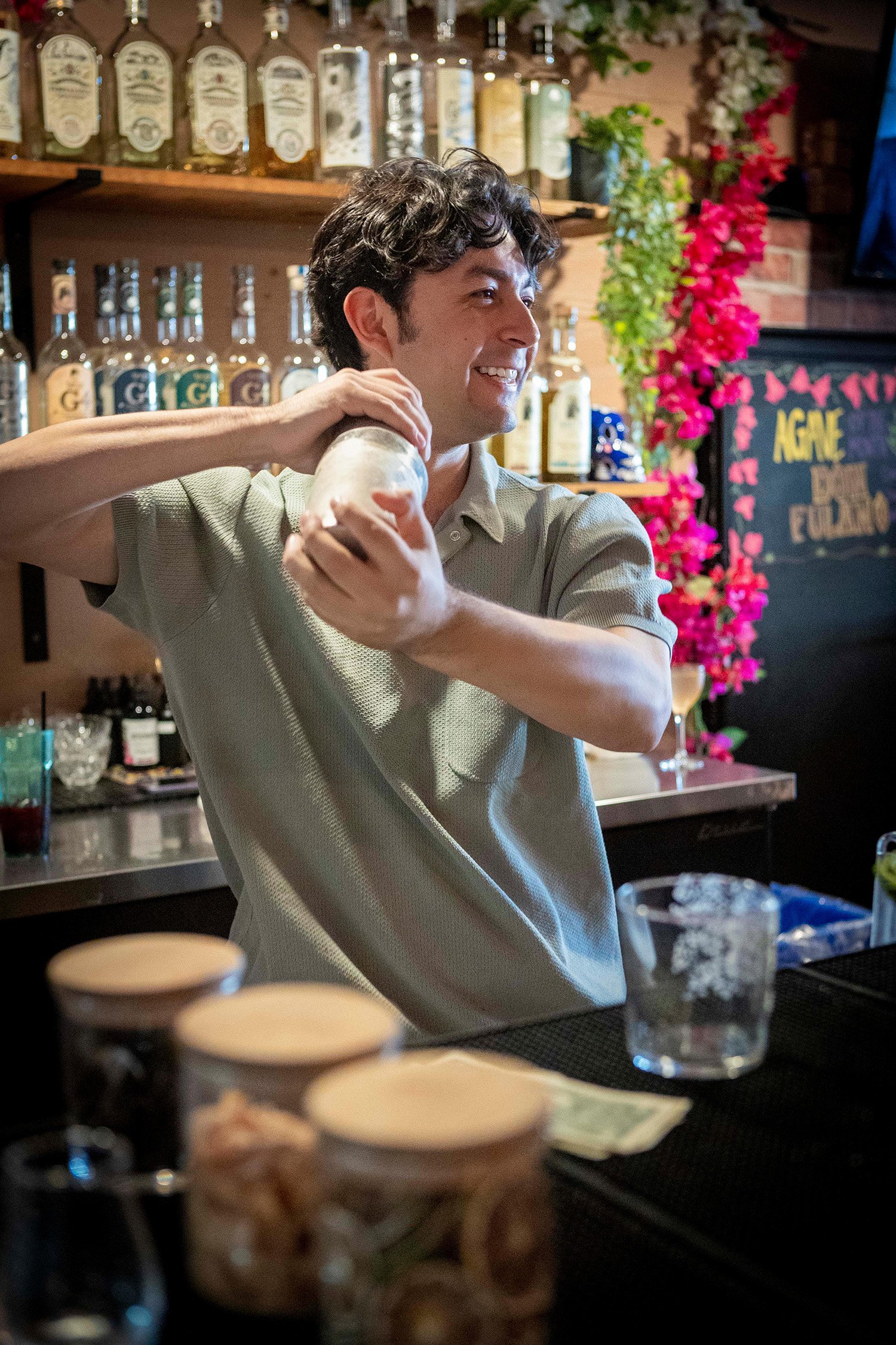 A man shaking a cocktail shaker behind a bar with alcohol bottles on the shelf and flowers in the background.