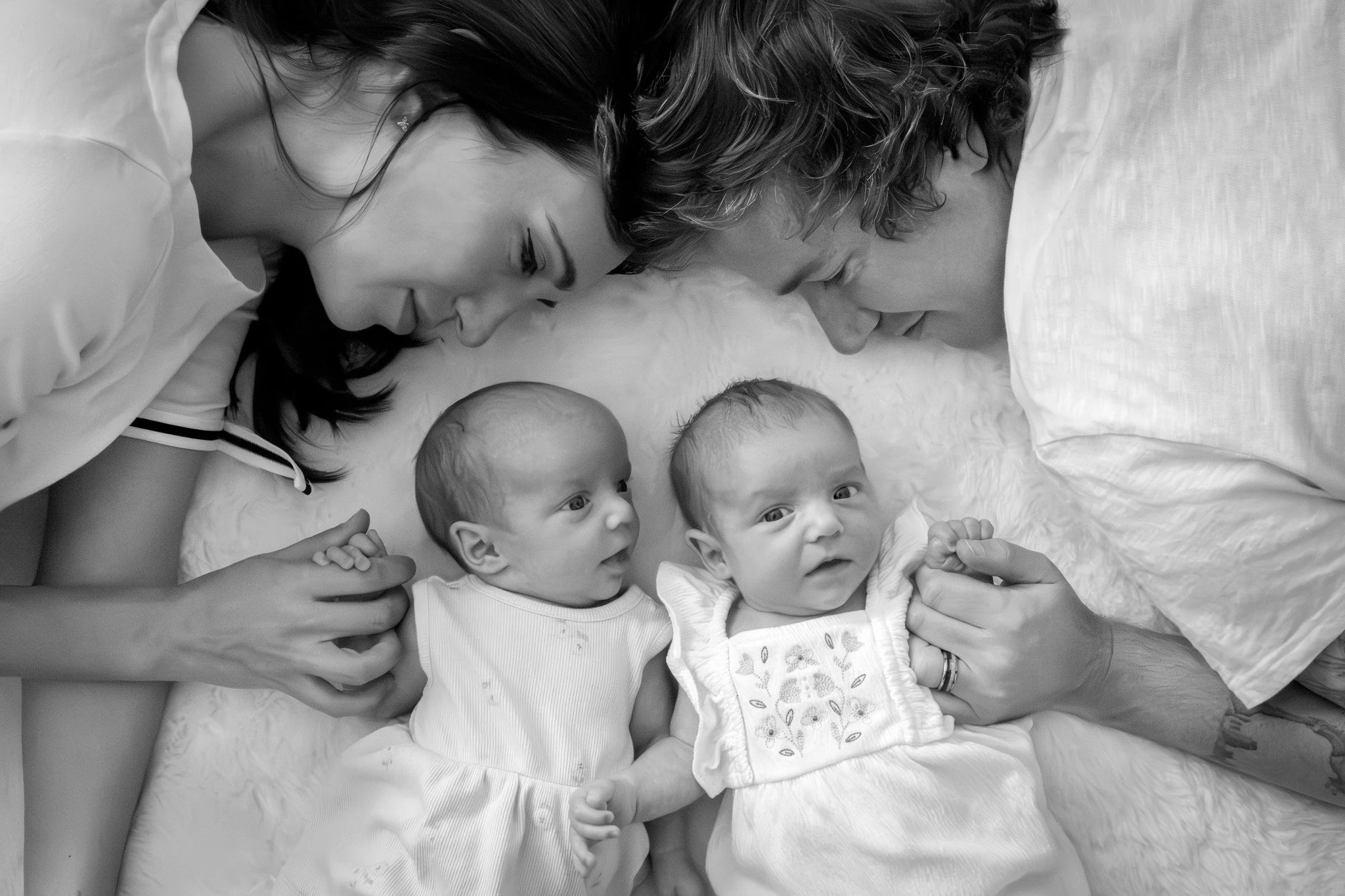 Black and white photo of two women and two babies lying on a fluffy surface, nose to nose, looking at each other, with one woman gently holding a baby's hand.