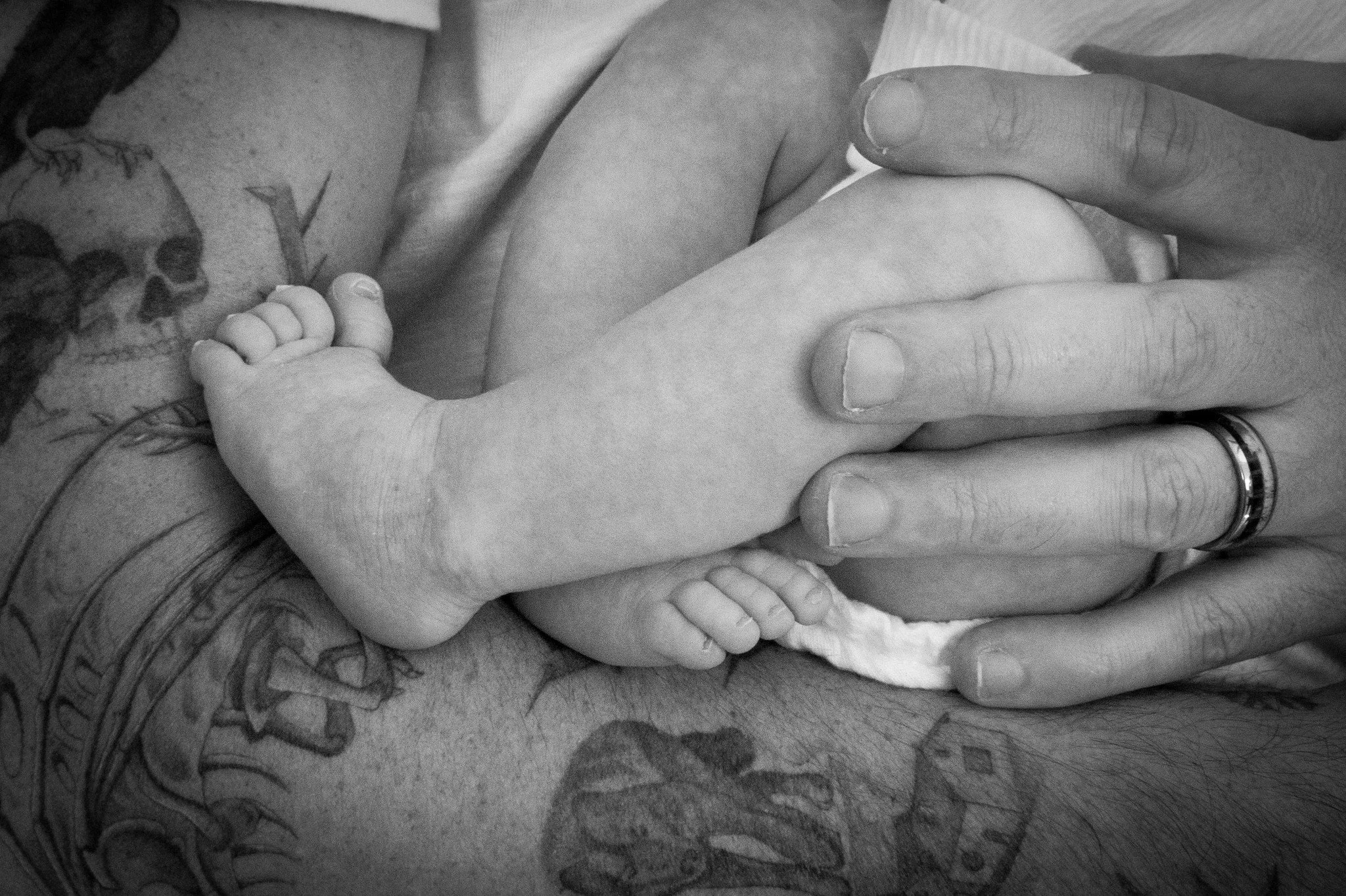 A close-up black and white photo of a person with tattoos on their arm holding and supporting a tiny baby with tiny feet. The baby's head and body are not clearly visible.