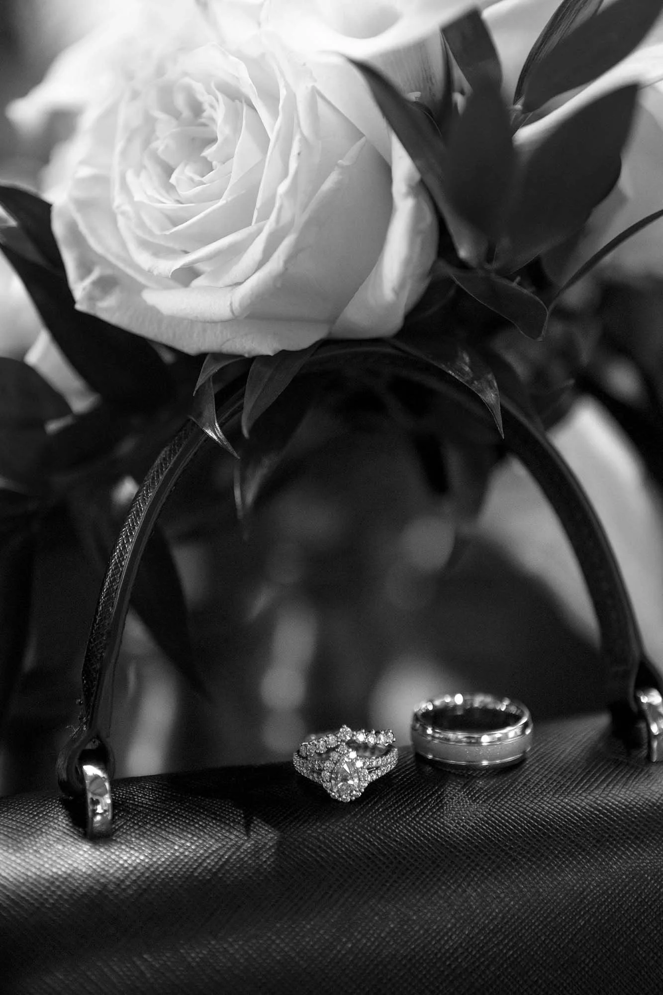 A black and white photo of a wedding bouquet with a large rose, two wedding rings, and a jeweled ring on a textured surface.