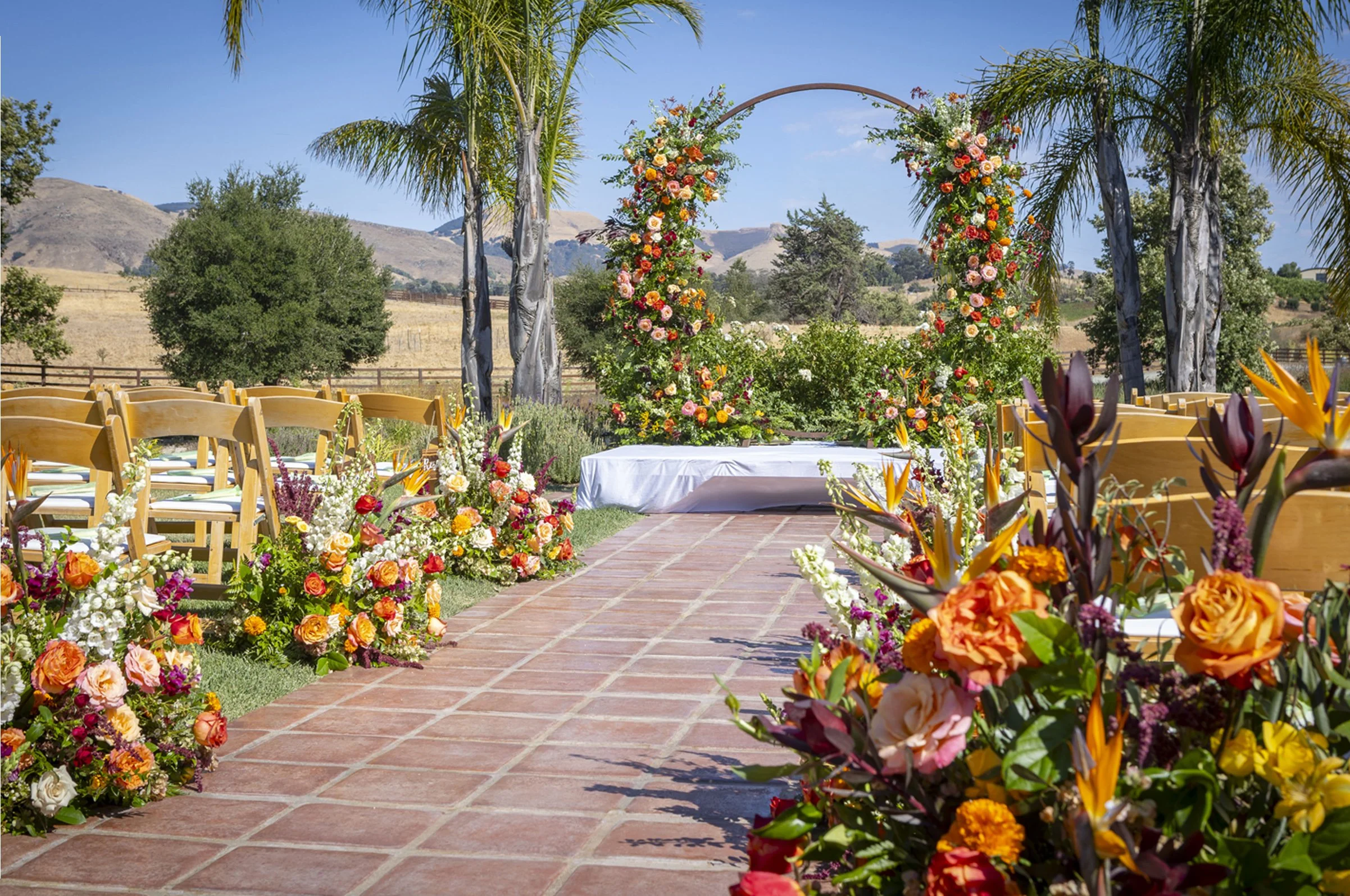 Outdoor wedding ceremony setup with wooden chairs, floral arrangements, and a floral arch, on a brick aisle with scenic hills and trees in the background.