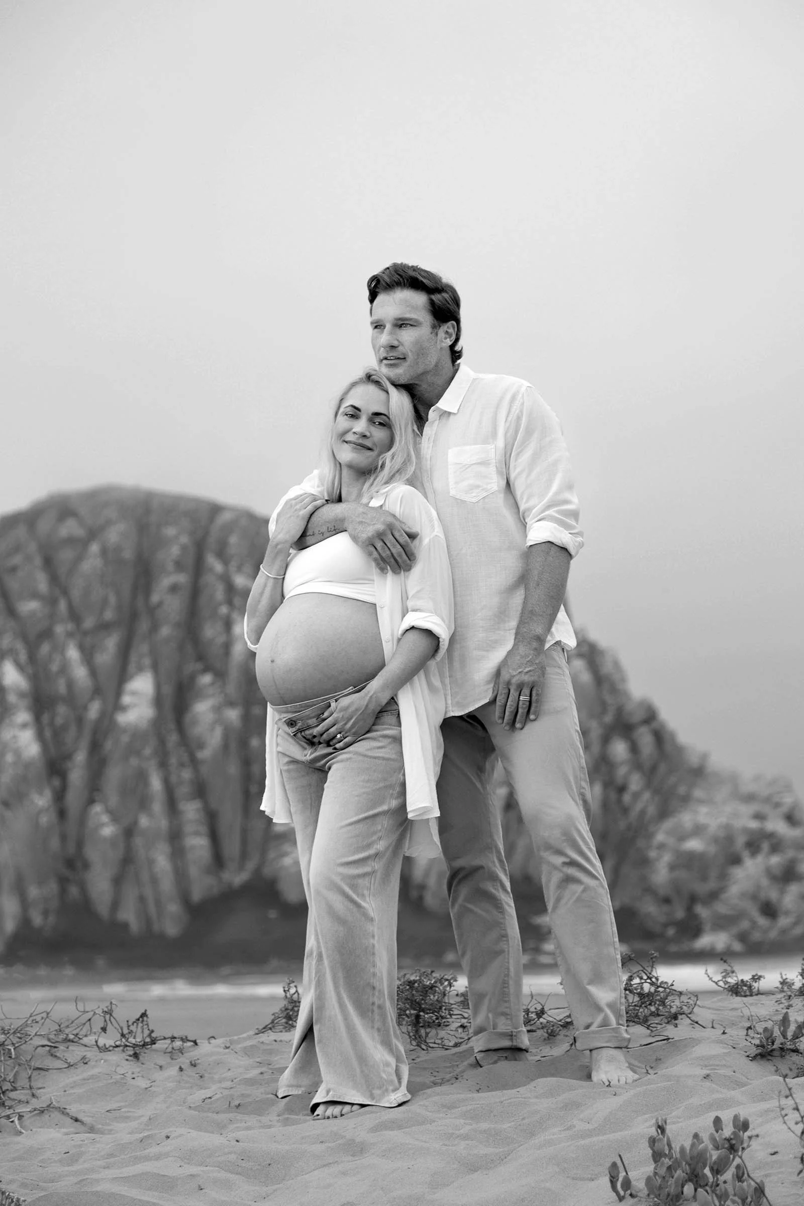 A pregnant woman and a man stand together on a sandy landscape with sparse vegetation, with trees in the background. The woman is smiling and gently touching her belly, while the man stands close with his arm around her shoulders, both dressed in cas