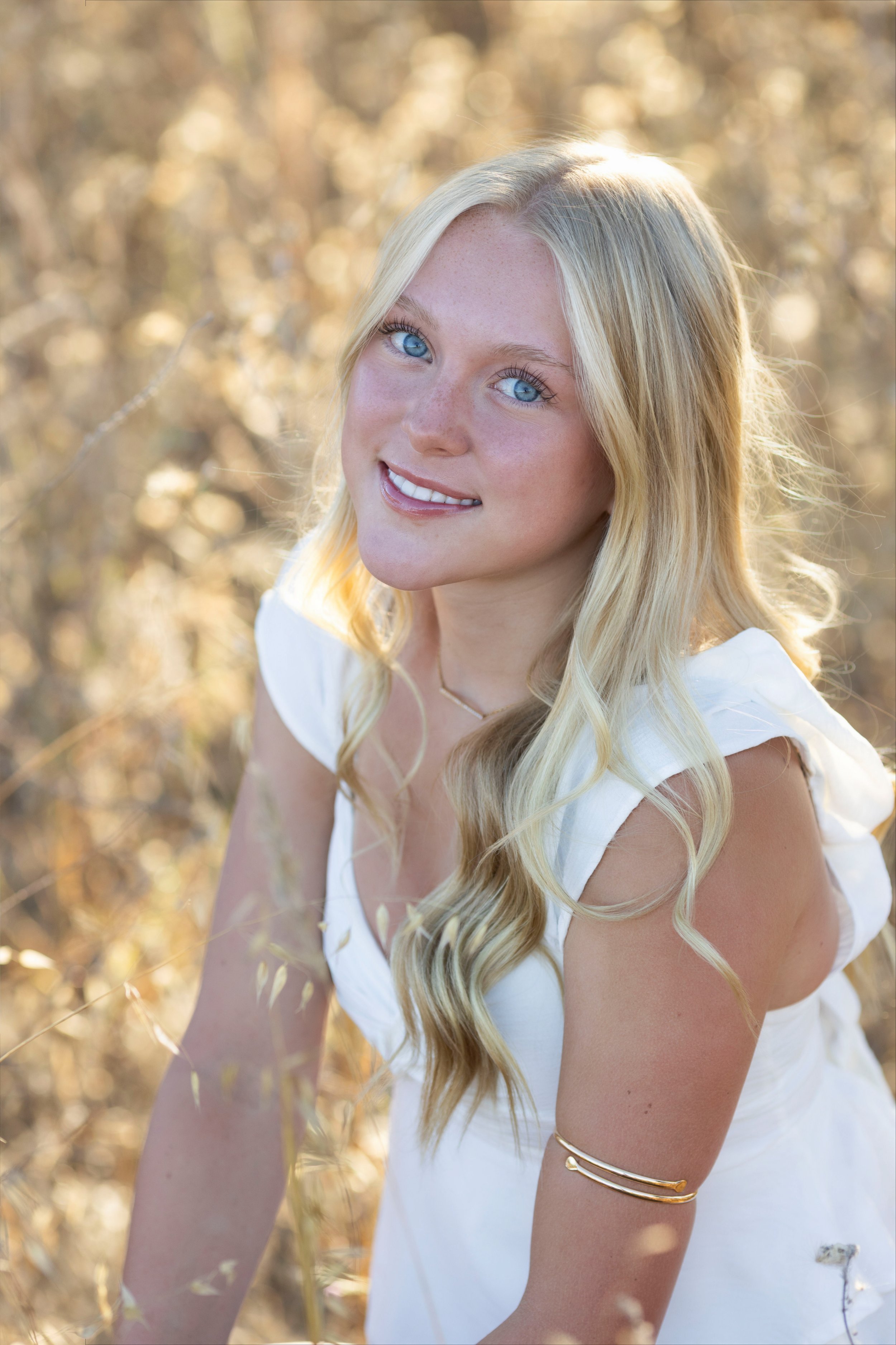 A young woman with long blonde hair and blue eyes smiling outdoors in a field with sunlight filtering through the trees.
