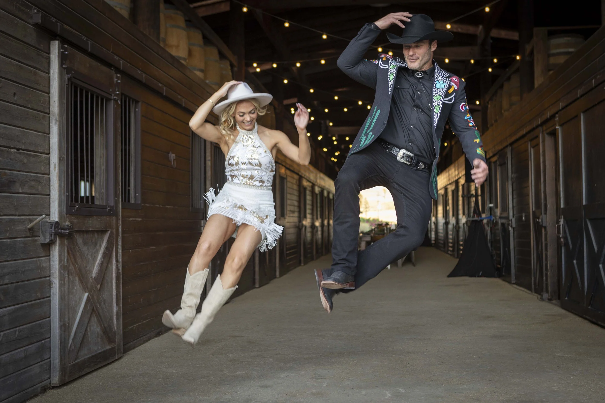 A man and woman wearing cowboy hats and Western-themed outfits dancing and jumping inside a barn with wooden walls and string lights.