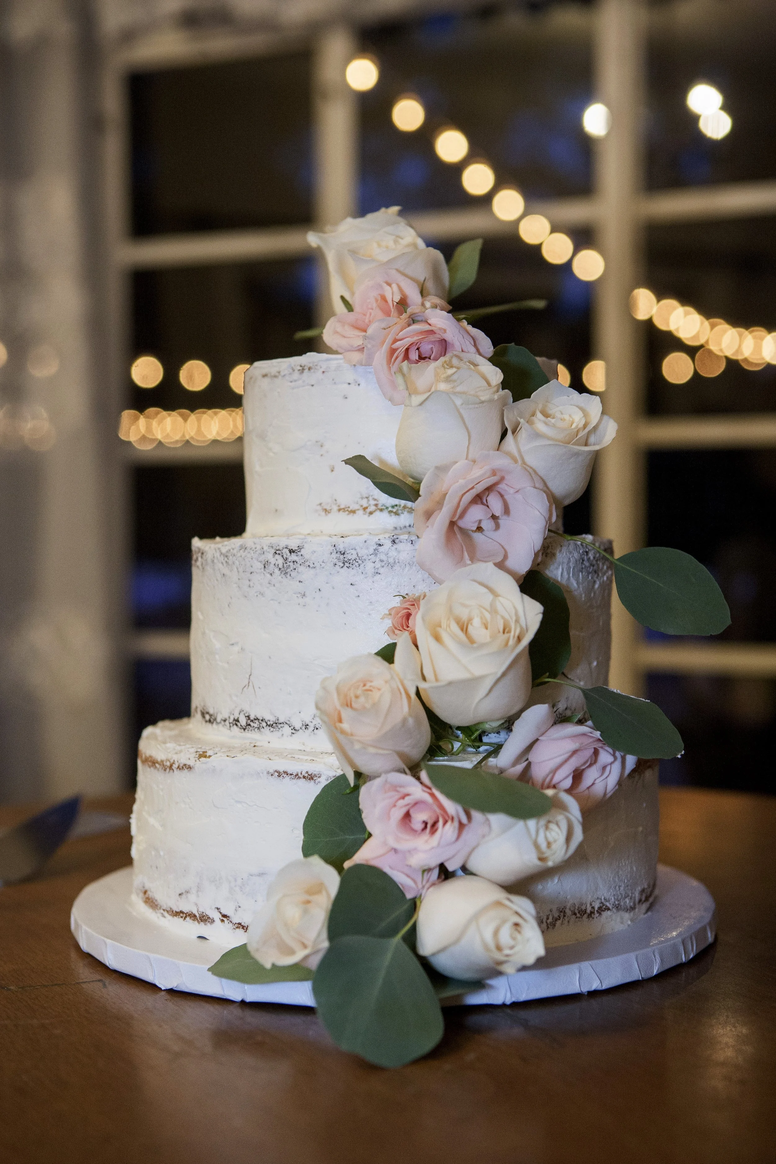 A three-tiered wedding cake decorated with white and light pink roses with green leaves, set on a wooden table with soft lighting and blurred warm string lights in the background.