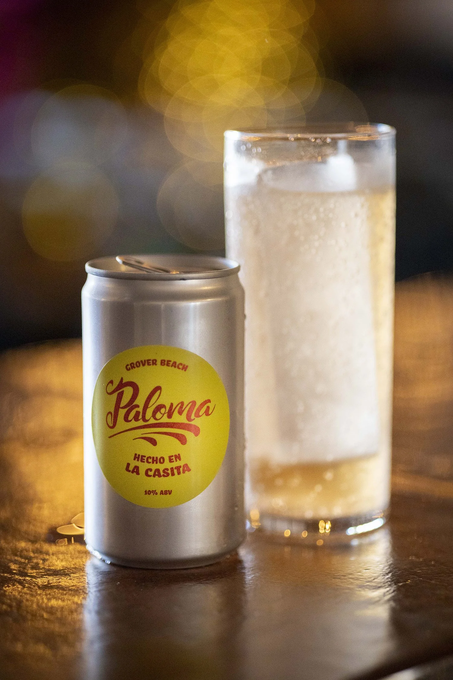 A canned beverage labeled 'Paloma' with a yellow circle and red text, alongside a tall glass of a pale, fizzy drink with ice cubes, on a wooden surface with blurred colorful lights in the background.