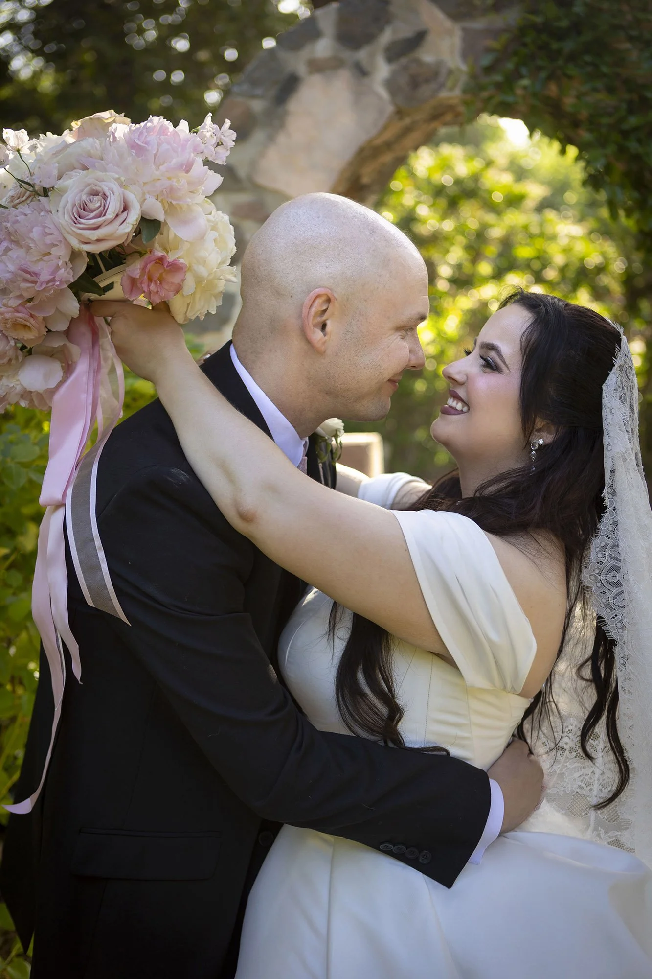 A bride and groom embrace, looking into each other's eyes, with the bride holding a bouquet of pink and white flowers. The bride has dark hair and is wearing a white gown and lace veil, while the groom is bald, wearing a black suit, outdoors with gre