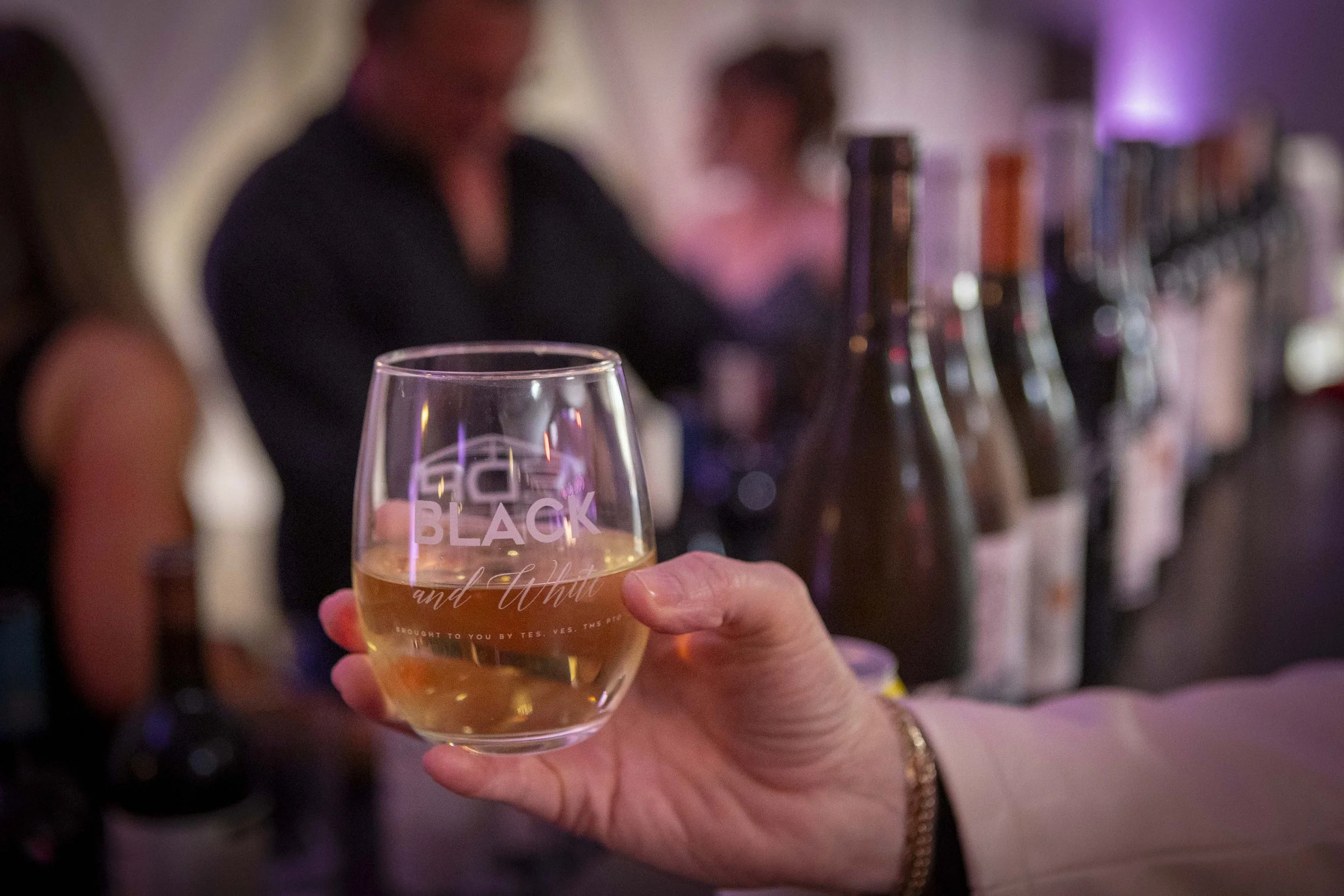 A hand holding a glass of white wine at a bar, with bottles lined up in the background, and bar patrons in a dimly lit environment.