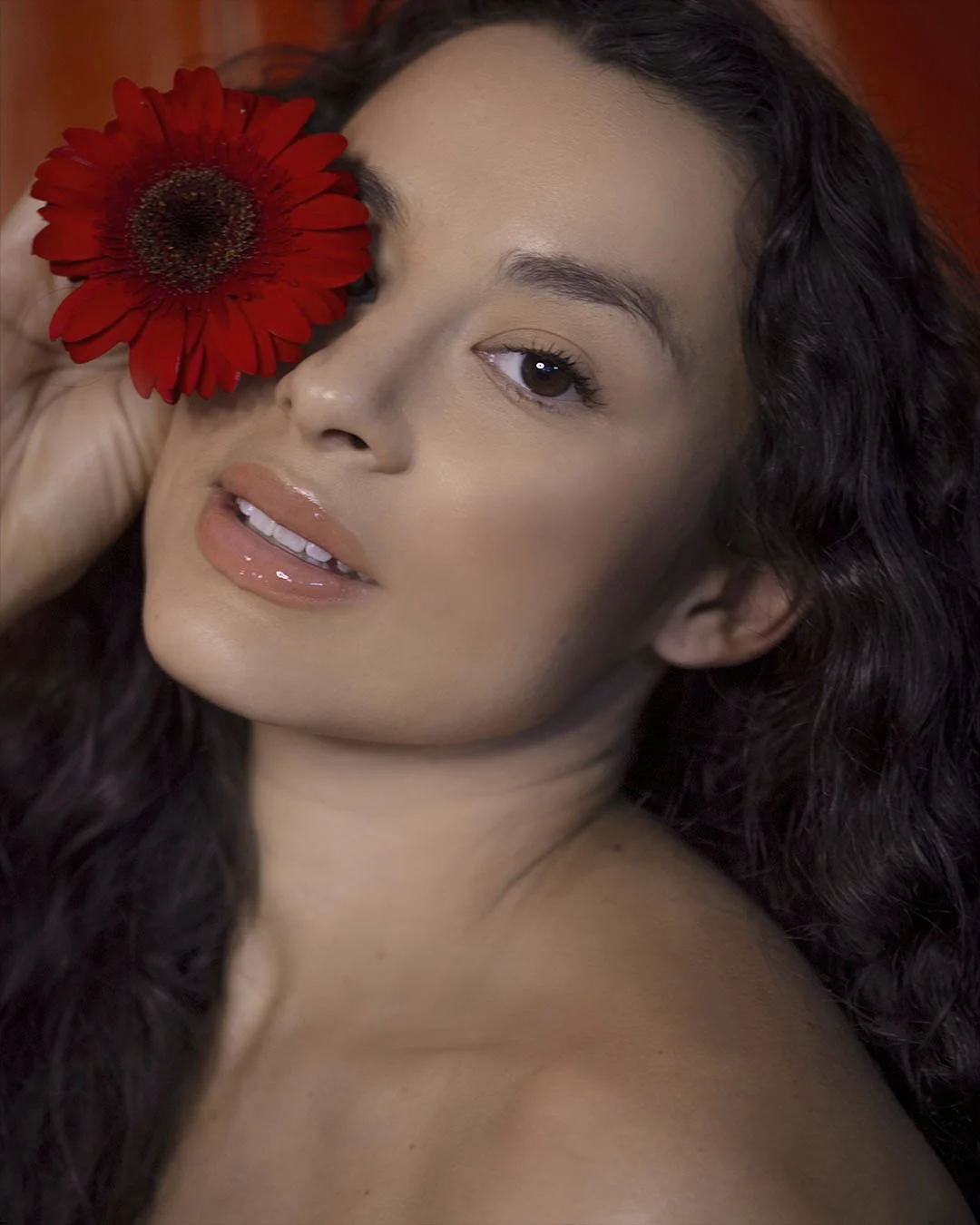 A woman with dark, curly hair holds a red flower over her left eye, looking directly at the camera with a subtle smile.