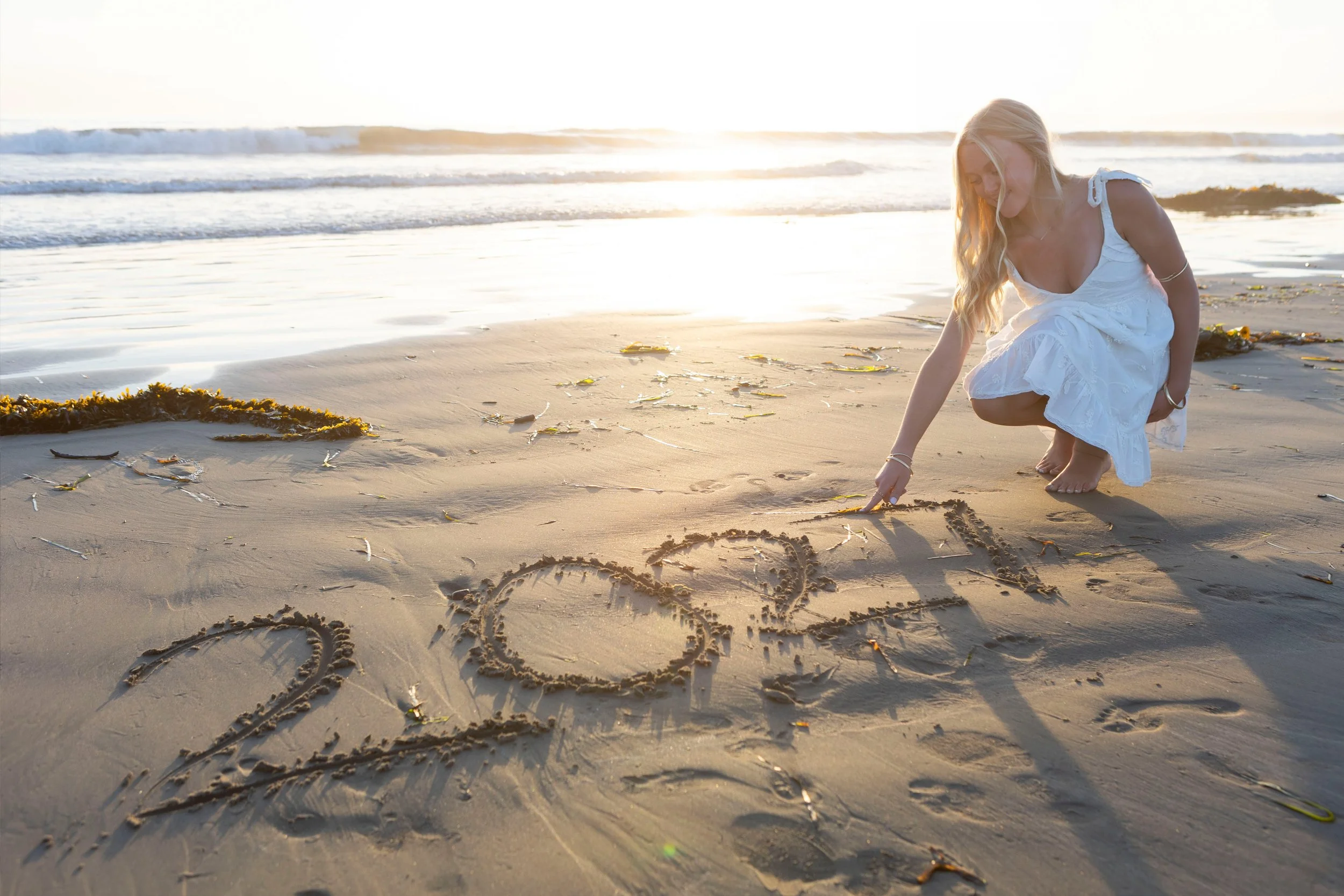A woman crouching on a sandy beach drawing in the sand with her finger as the sun sets or rises over the ocean, with waves and seaweed in the background.