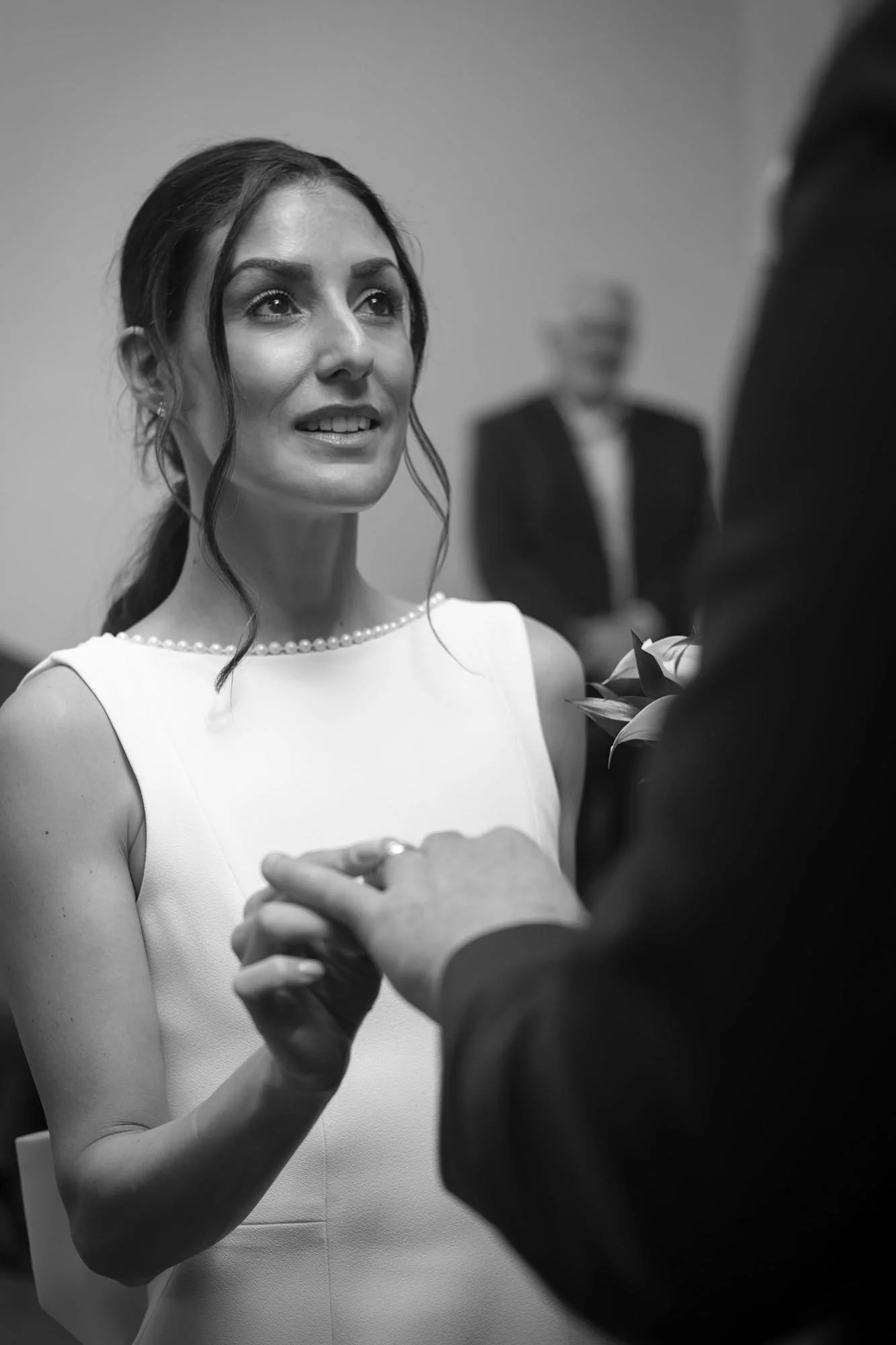 A black-and-white photo of a woman during a wedding ceremony, holding hands with a man. The woman has dark hair styled in loose waves, is wearing a sleeveless dress with a pearl neckline, and has a gentle expression as she looks at the man. The man i