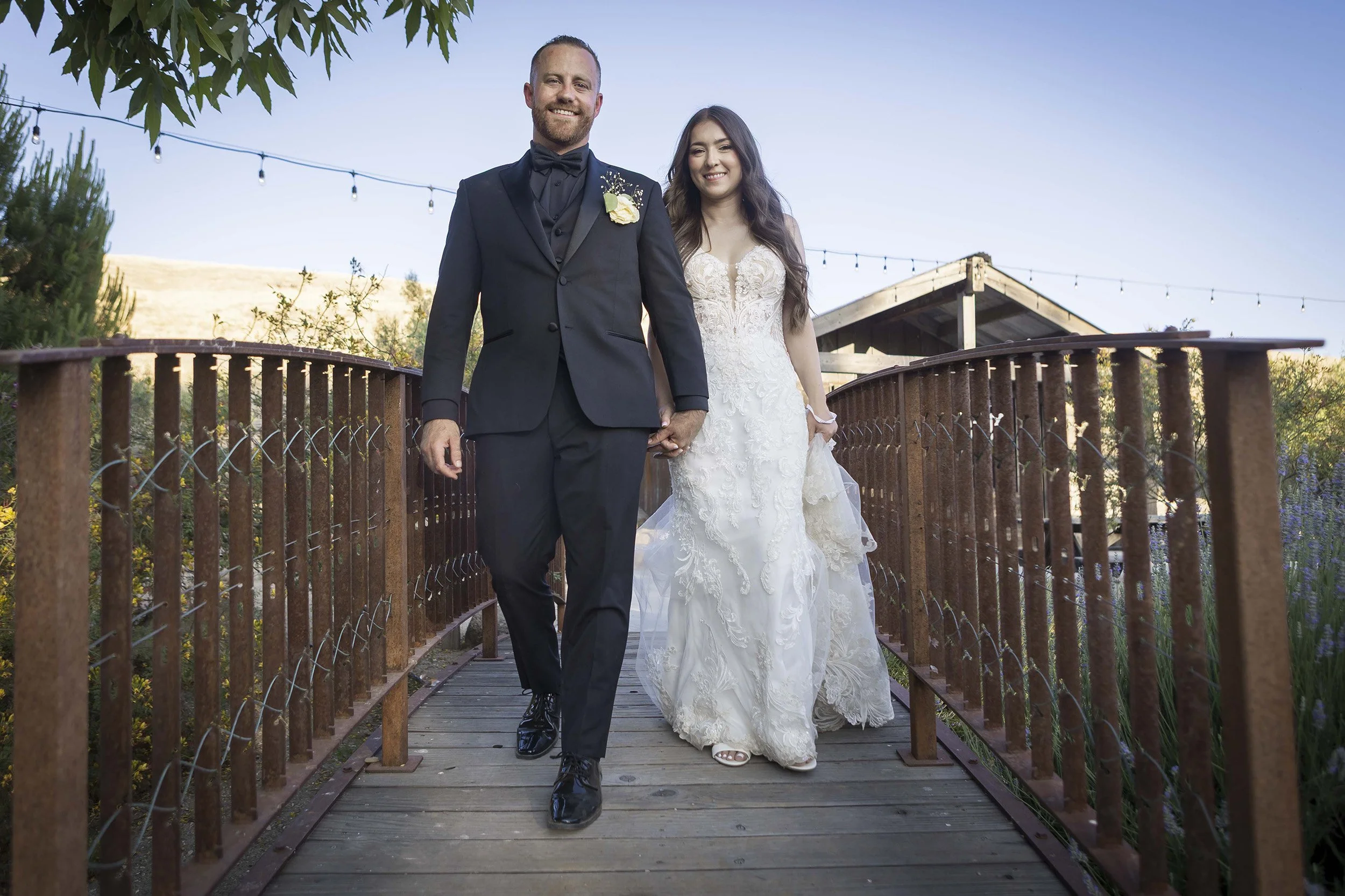 A newlywed couple walking hand-in-hand on a wooden bridge outdoors, surrounded by greenery with string lights above them, during daytime.