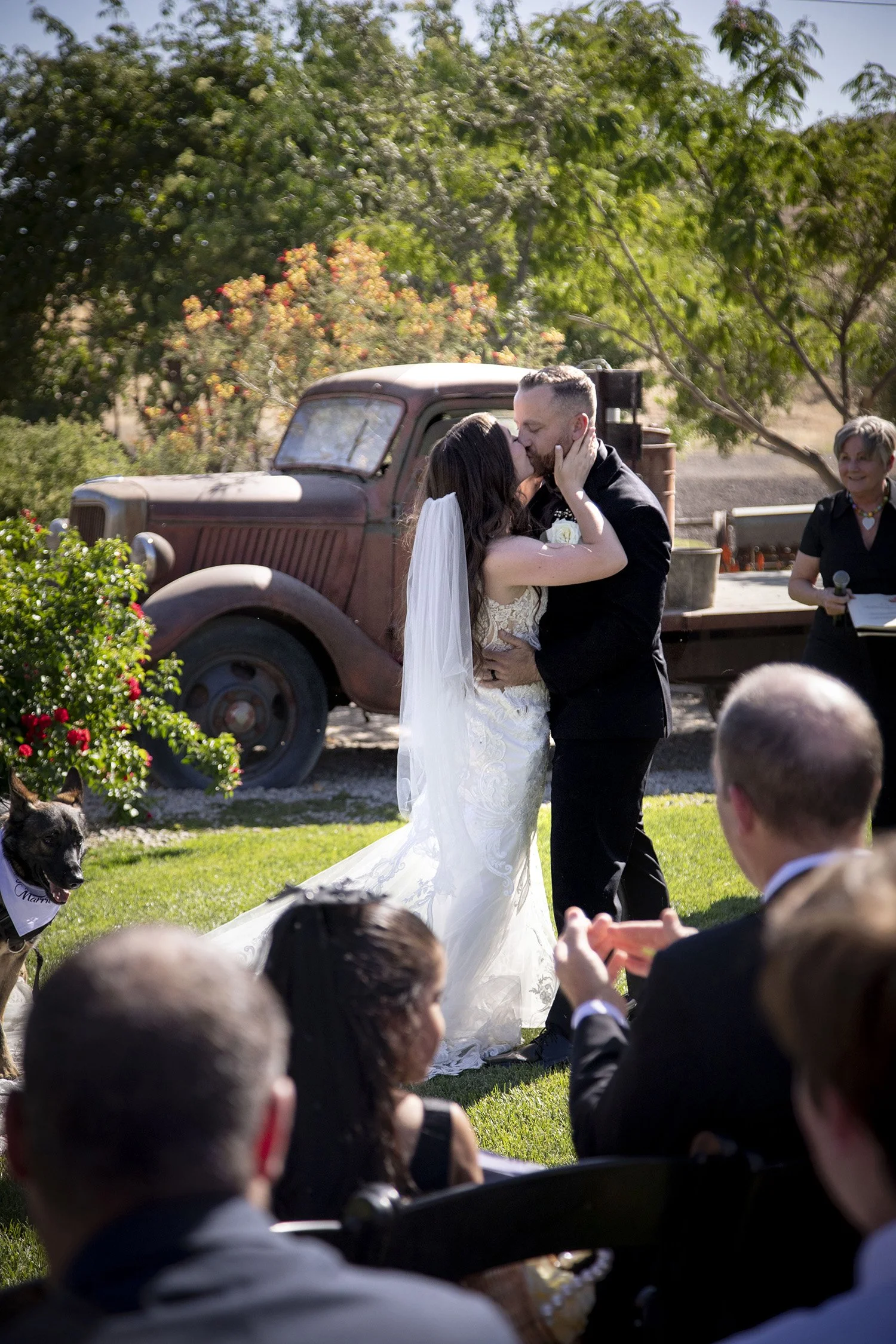 A couple shares a kiss during a wedding ceremony outdoors, with guests seated around them and a vintage truck in the background.