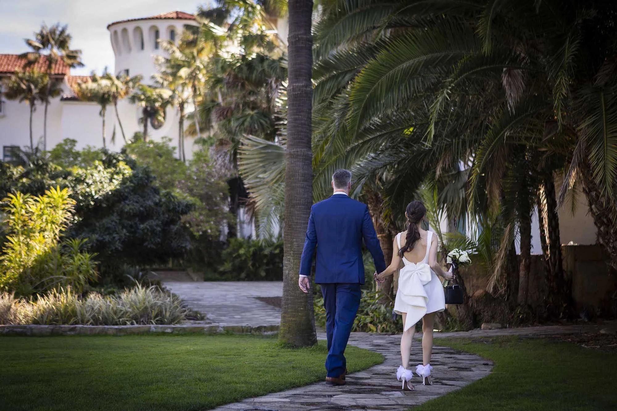 A couple dressed in wedding attire walking on a stone pathway through a lush tropical garden with palm trees, shrubs, and a white building with a tiled roof in the background.