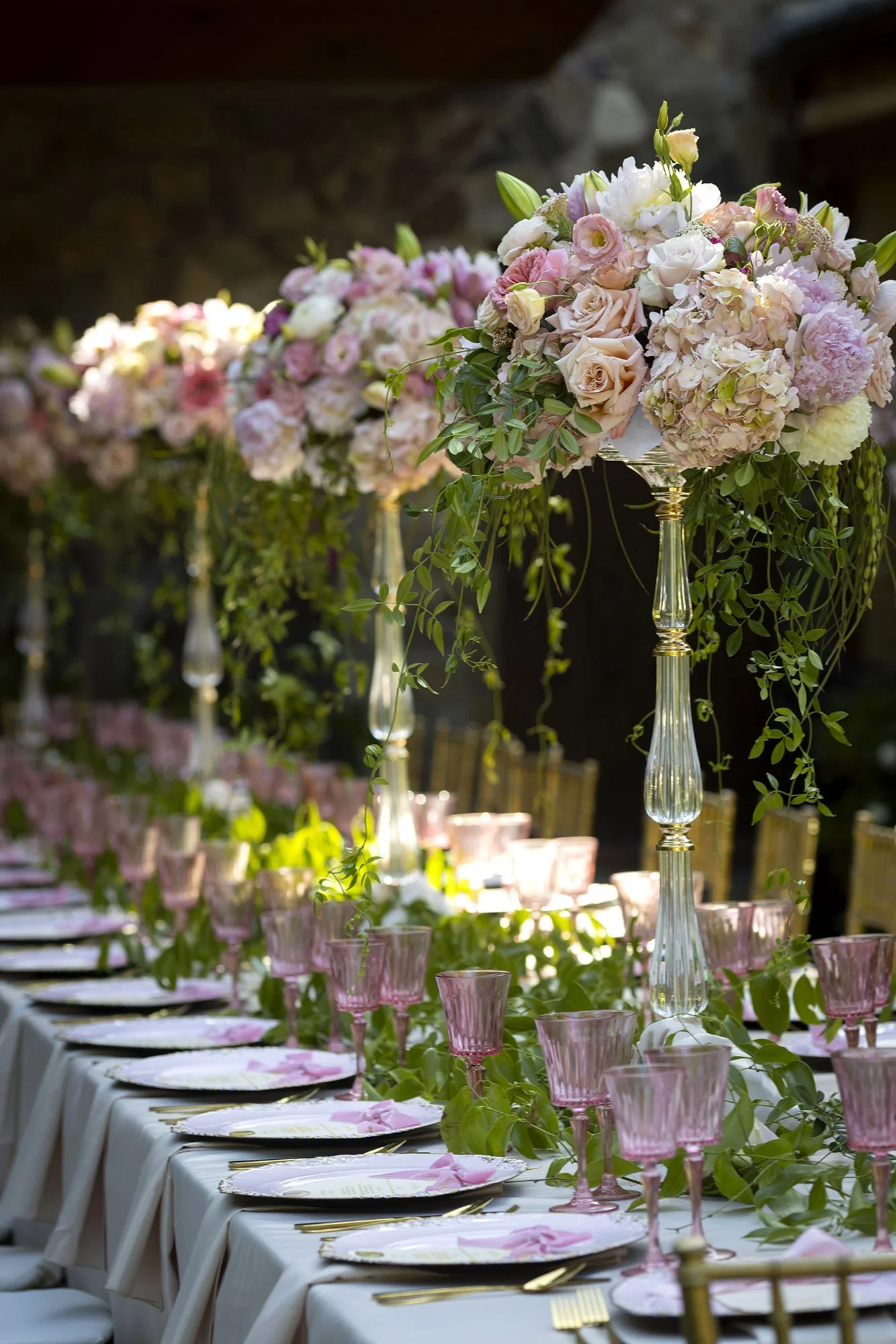 A long banquet table decorated with tall floral centerpieces featuring pink and white flowers and hanging green vines, set with pink glasses, gold cutlery, and floral napkin accents for a celebration.
