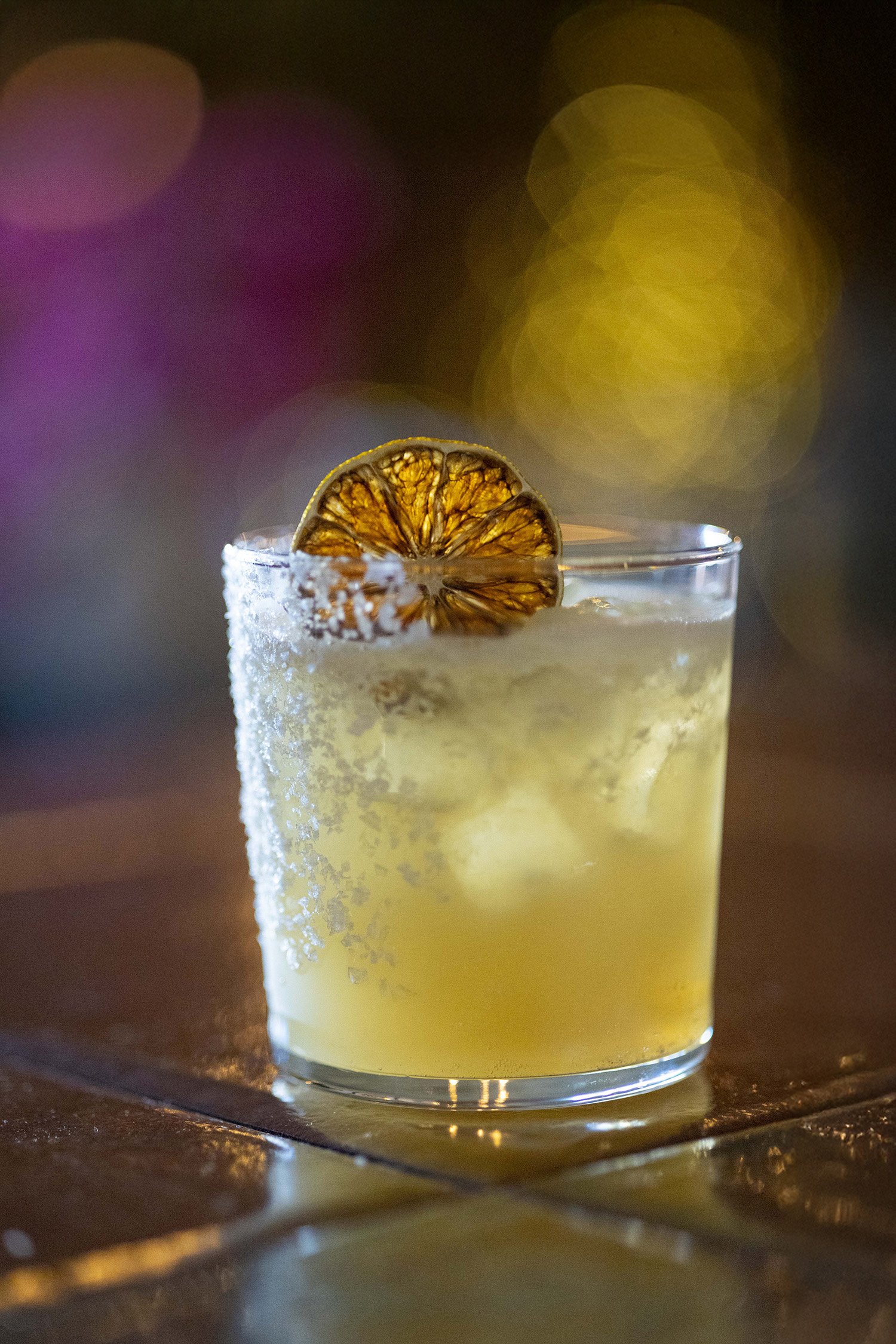 A glass of cocktail garnished with a dried citrus slice, with salted rim and ice cubes inside.
