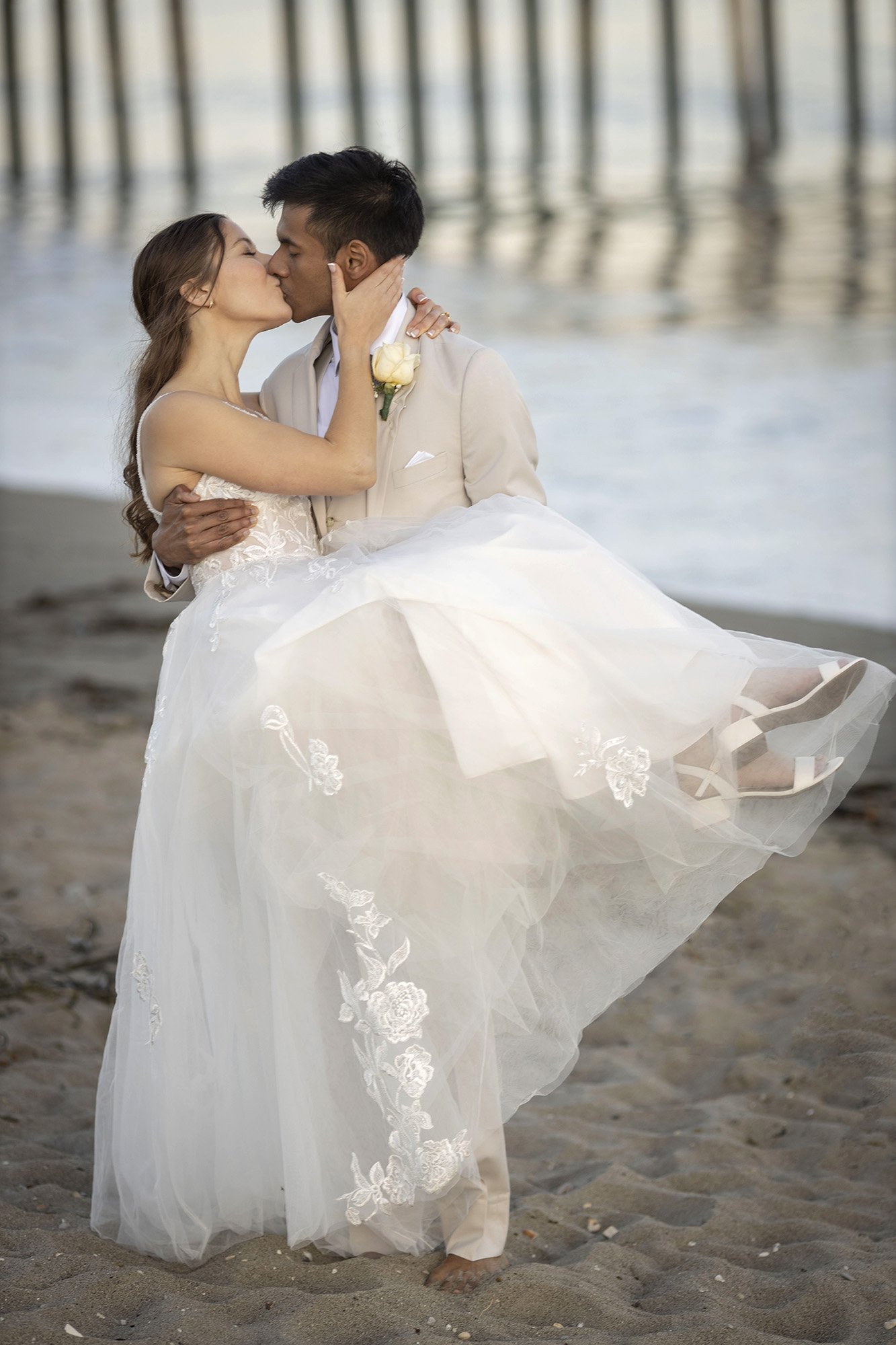 A bride and groom sharing a kiss on the beach, with the groom carrying the bride in his arms as they stand on sand with water and a pier in the background.