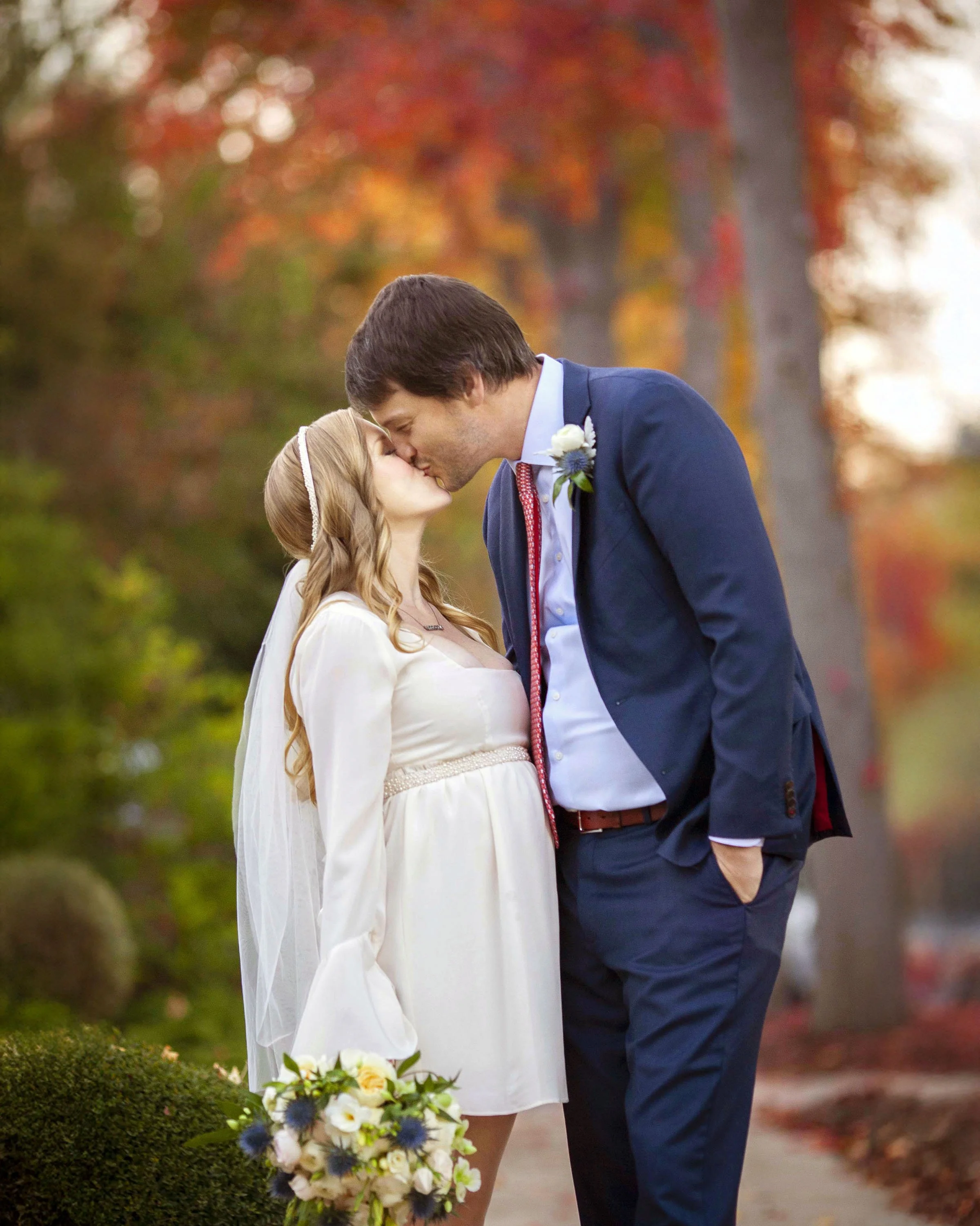 A couple sharing a kiss outdoors with autumn trees in the background, the woman in a white dress holding a bouquet of flowers, the man in a navy suit with a boutonniere.