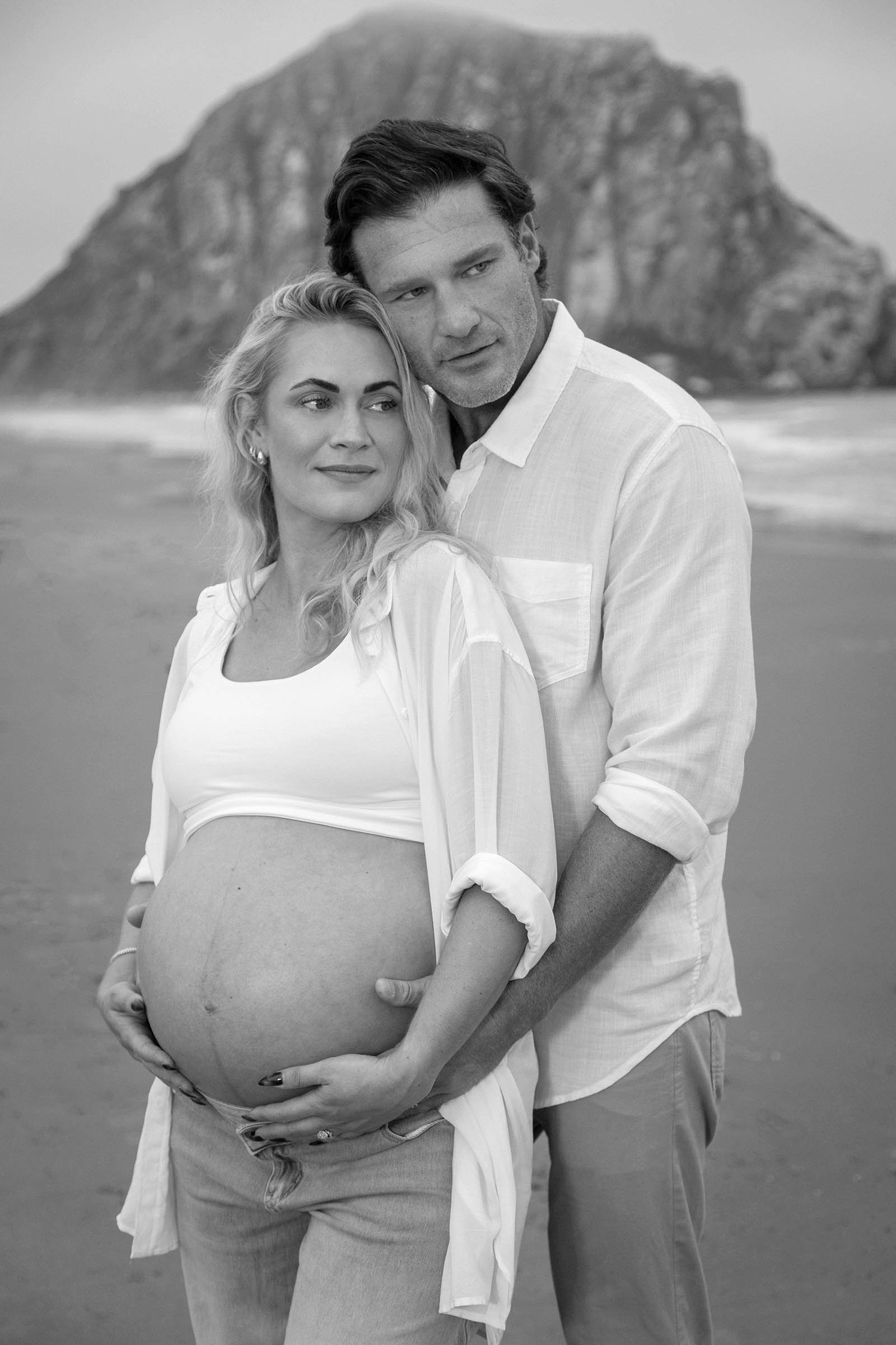 Black and white photo of a couple at the beach, with the woman visibly pregnant. She is smiling softly, wearing a white crop top and jeans, while the man, standing behind her, looks into the distance. A large mountain or rock formation is in the back