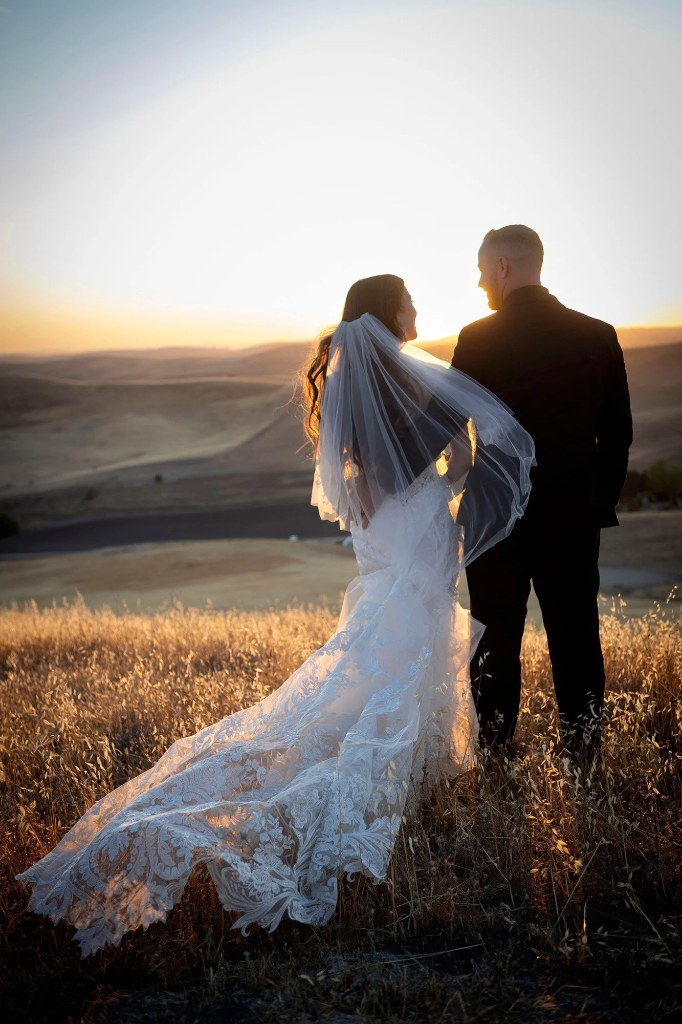 A bride and groom standing in a field during sunset, facing each other with the bride wearing a lace wedding gown and the groom in a black suit.