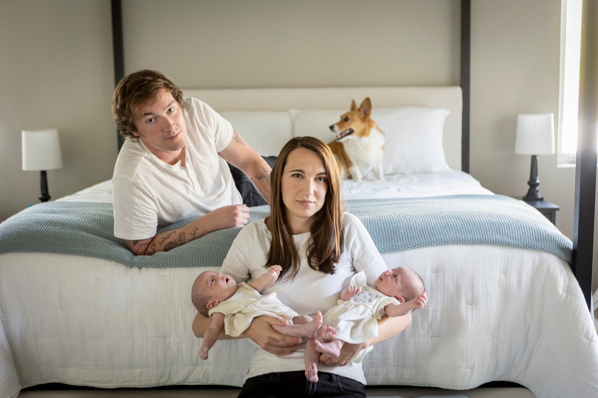 A woman sitting on the floor holding two babies, a man leaning on a bed with two dogs on it, in a bedroom with white walls and lamps.
