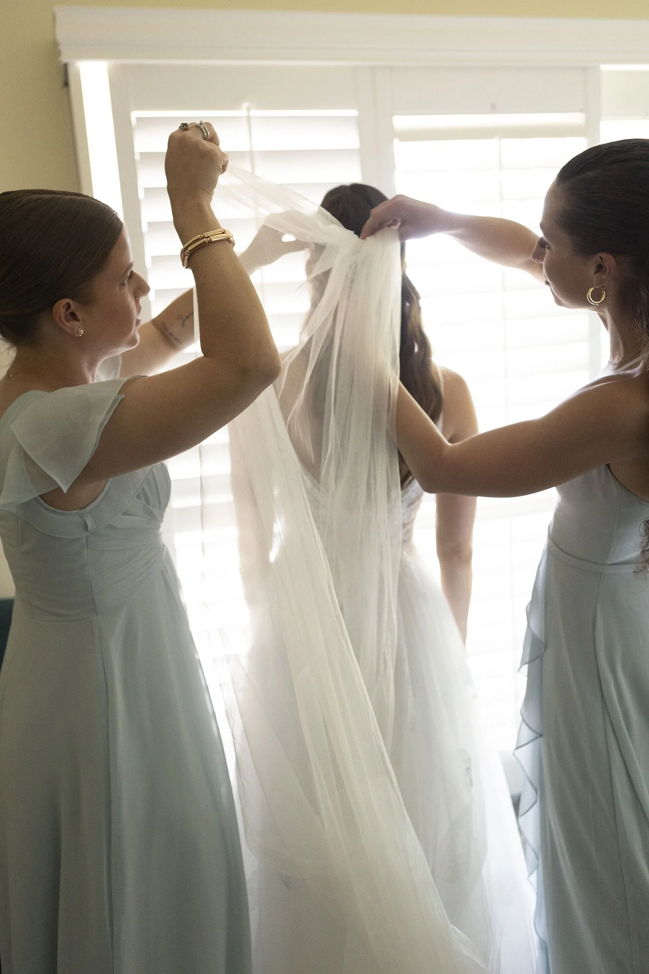 Women helping bride with wedding veil in front of window with blinds.