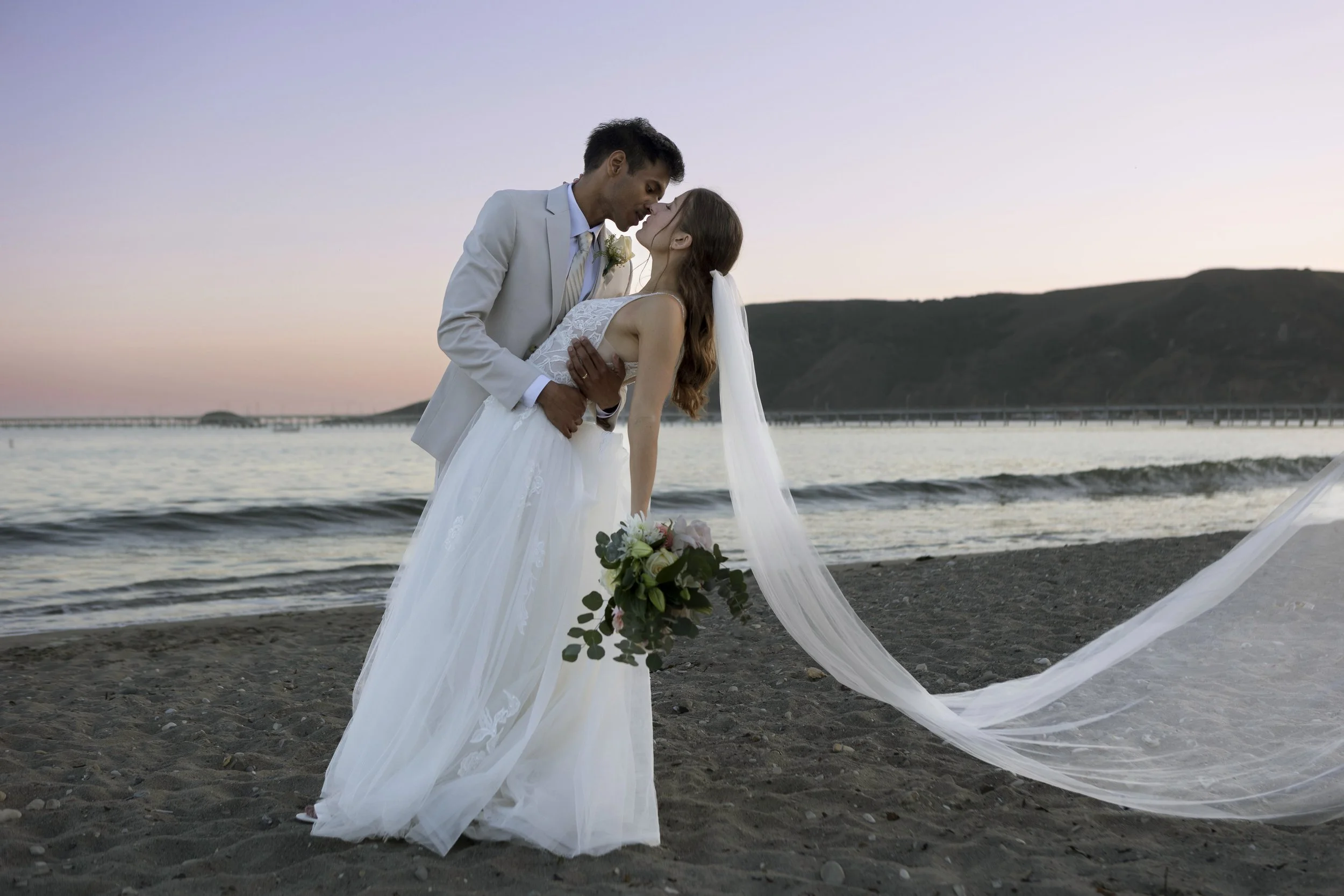 A newly married couple sharing a dance on the beach at sunset, the groom dressed in a light-colored suit and the bride in a white wedding gown with a veil, holding a bouquet of flowers.