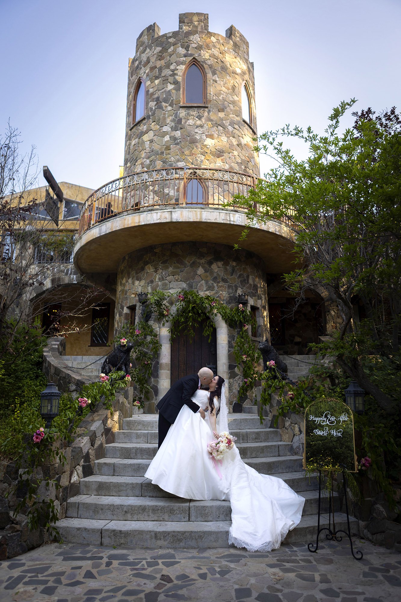 A newlywed couple sharing a kiss on the steps in front of a stone castle-like building, with a sign that says 'Happily Ever After Starts Here.' The bride is holding a bouquet of flowers, and there are decorative lions and flowers along the staircase.