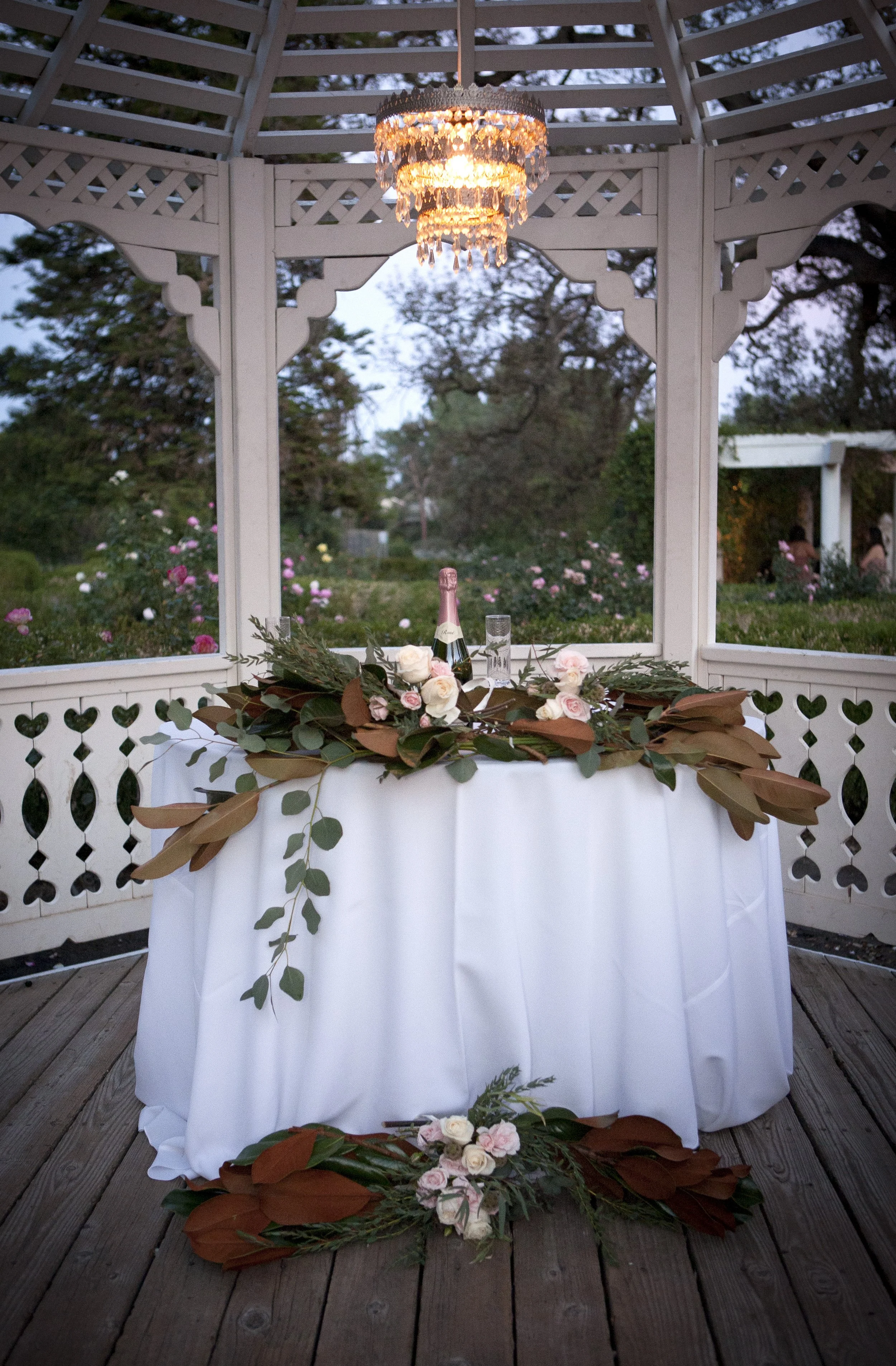 Decorative outdoor table with white tablecloth, floral arrangements, a champagne bottle, and glassware under a gazebo with a chandelier, overlooking a garden with blooming flowers.
