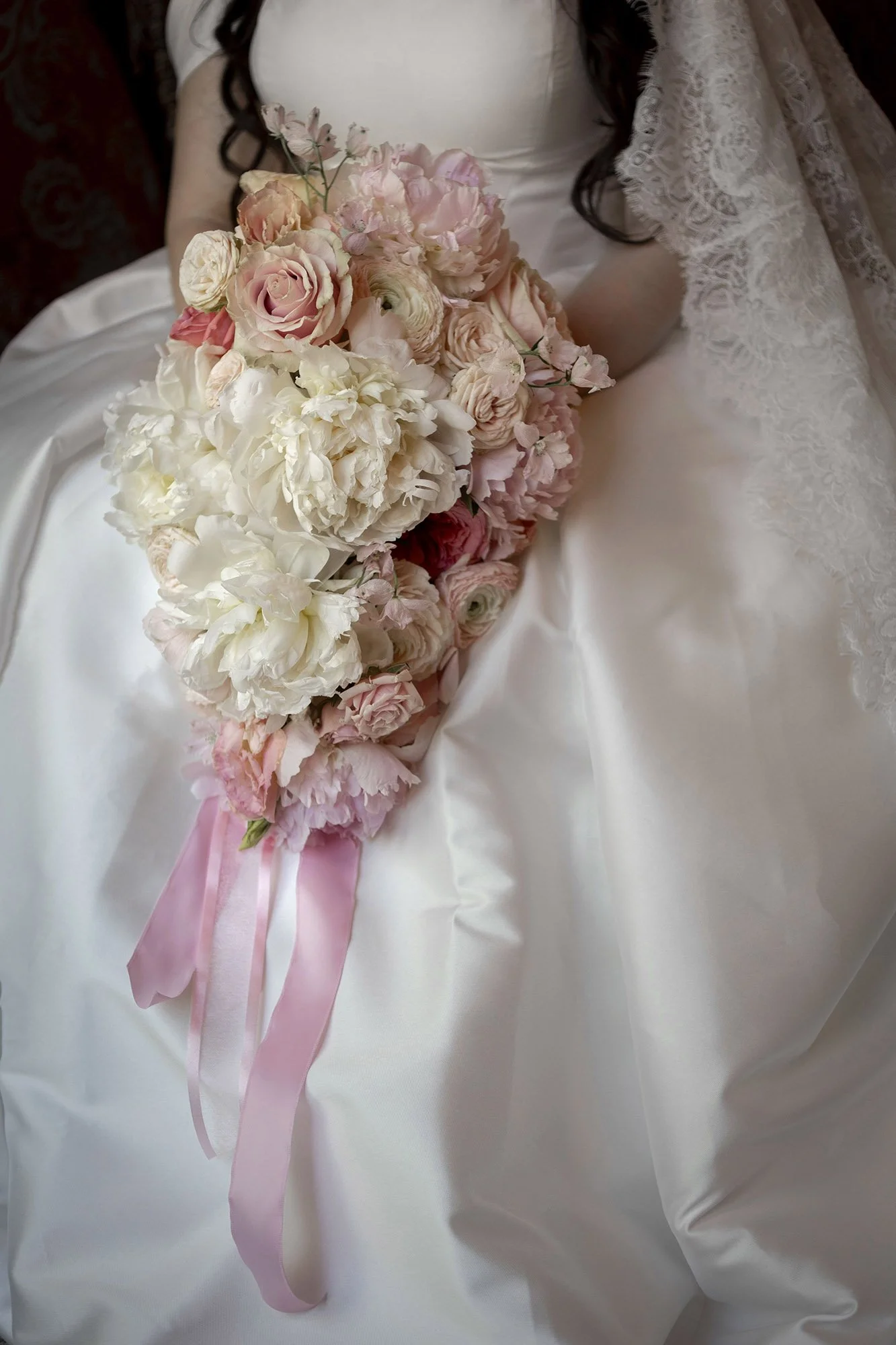 A bride in a white wedding gown holding a cascading bouquet of pink and white flowers, including roses and peonies, with pink ribbons.