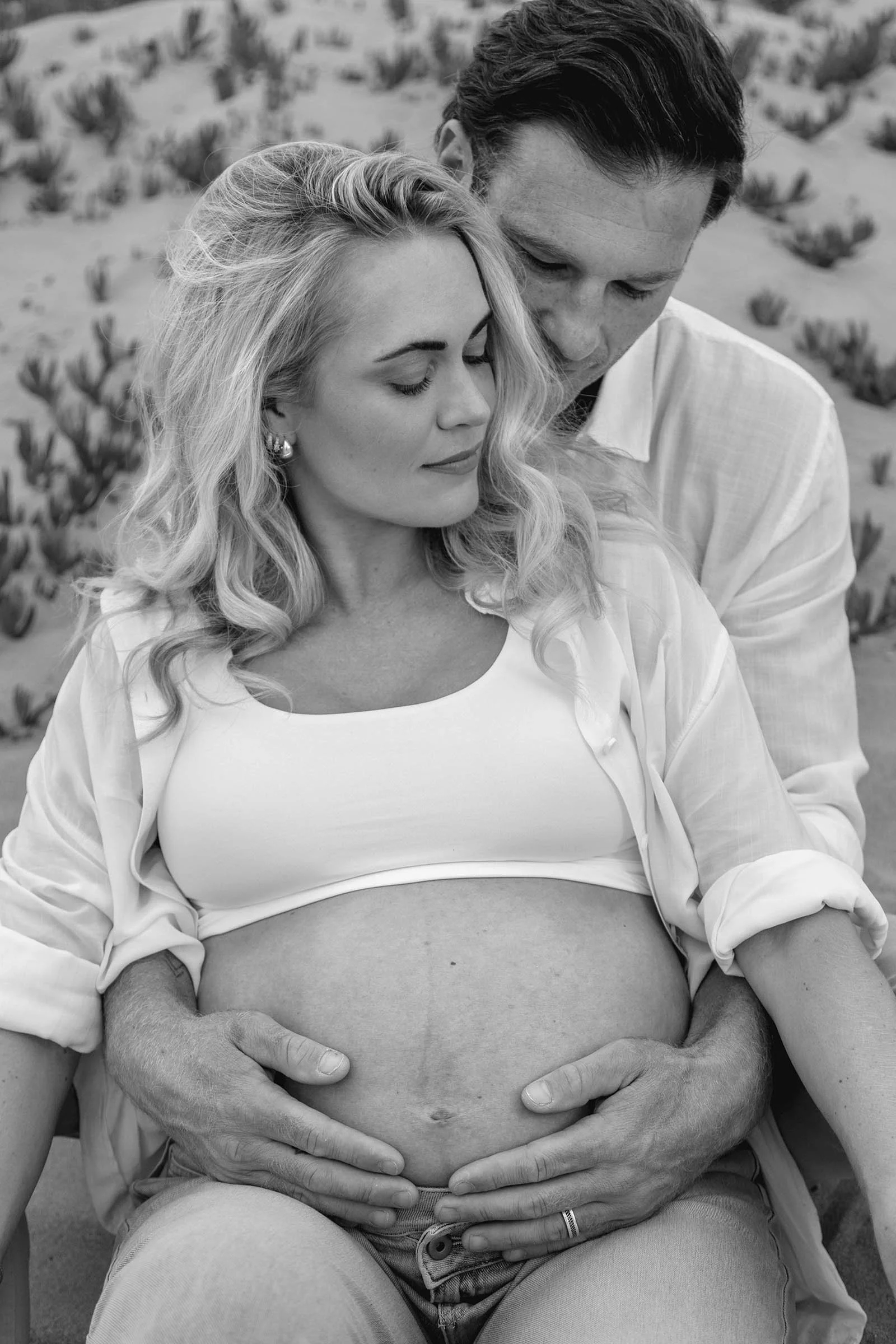 A pregnant woman with long blonde hair and earrings sits with closed eyes, her hands resting on her belly, while a man with dark hair, dressed in a light-colored shirt, embraces her from behind with his eyes also closed, against a background of deser