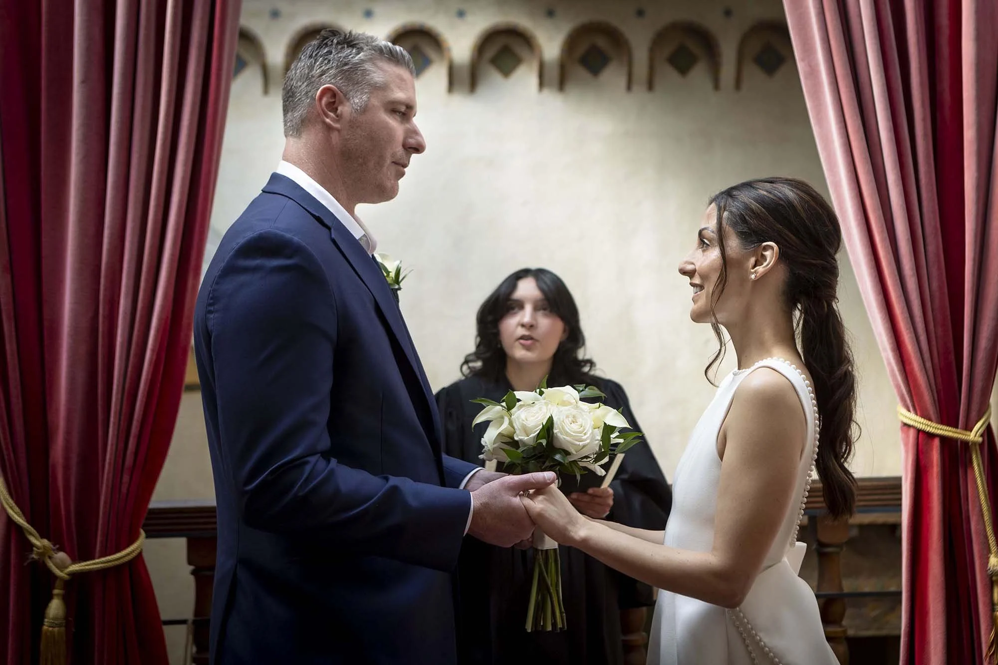 A couple getting married during their wedding ceremony, holding hands and exchanging vows, with an officiant behind them, seen through red curtains in an indoor setting.