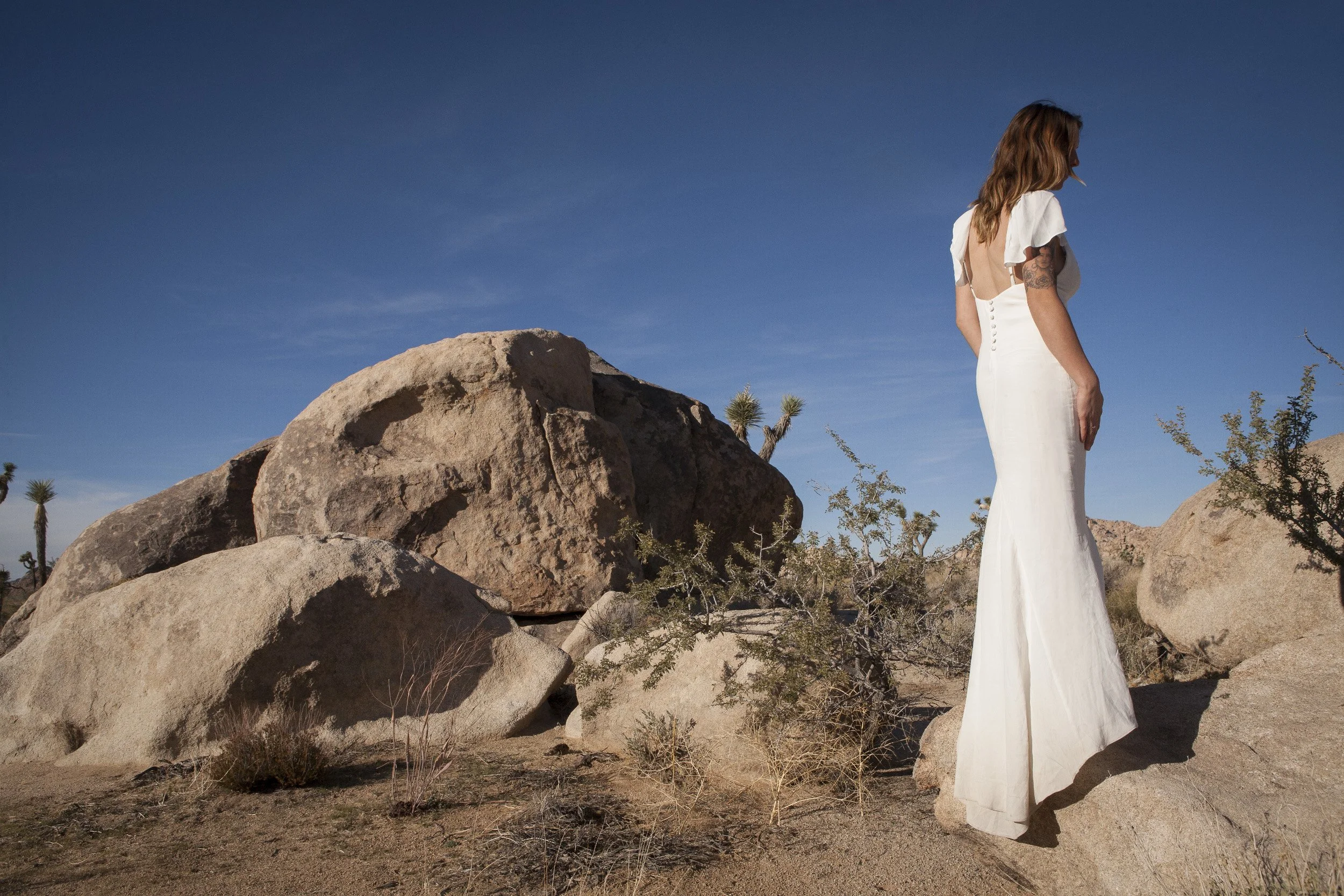 A woman in a white, backless dress standing on a rock in a desert landscape with large boulders and sparse vegetation under a blue sky.