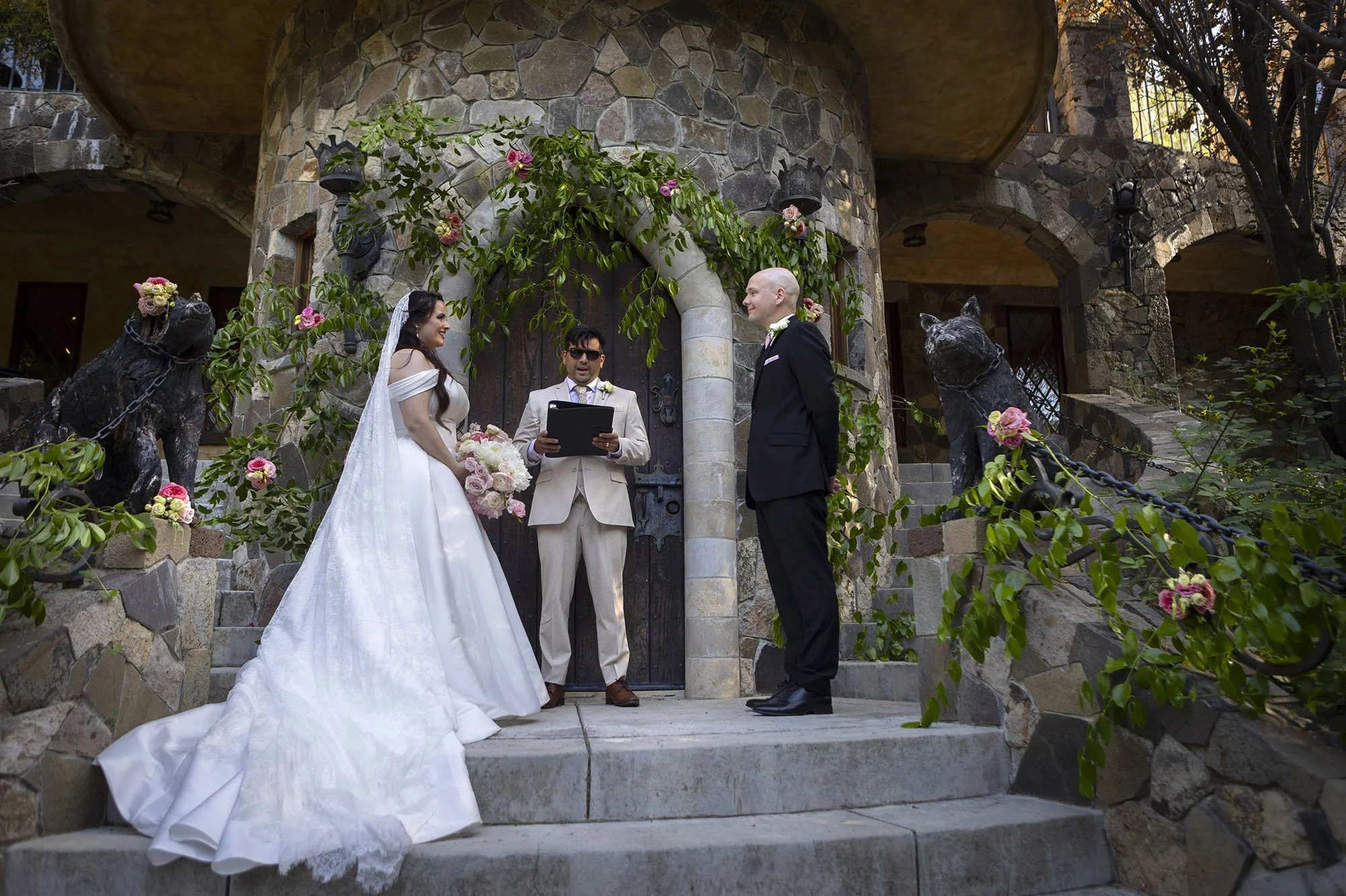 A wedding ceremony with a bride in a white wedding dress and veil, a groom in a black suit, and an officiant in a light beige suit, standing on stone steps outside a stone building decorated with flowers and greenery, with ornate statues of dogs.