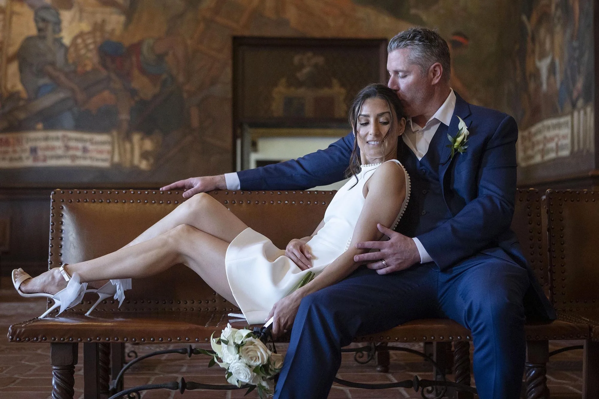 A newlywed couple sitting on a bench with the bride leaning back and smiling while the groom, in a navy suit, kisses her forehead in a decorated room with a mural.