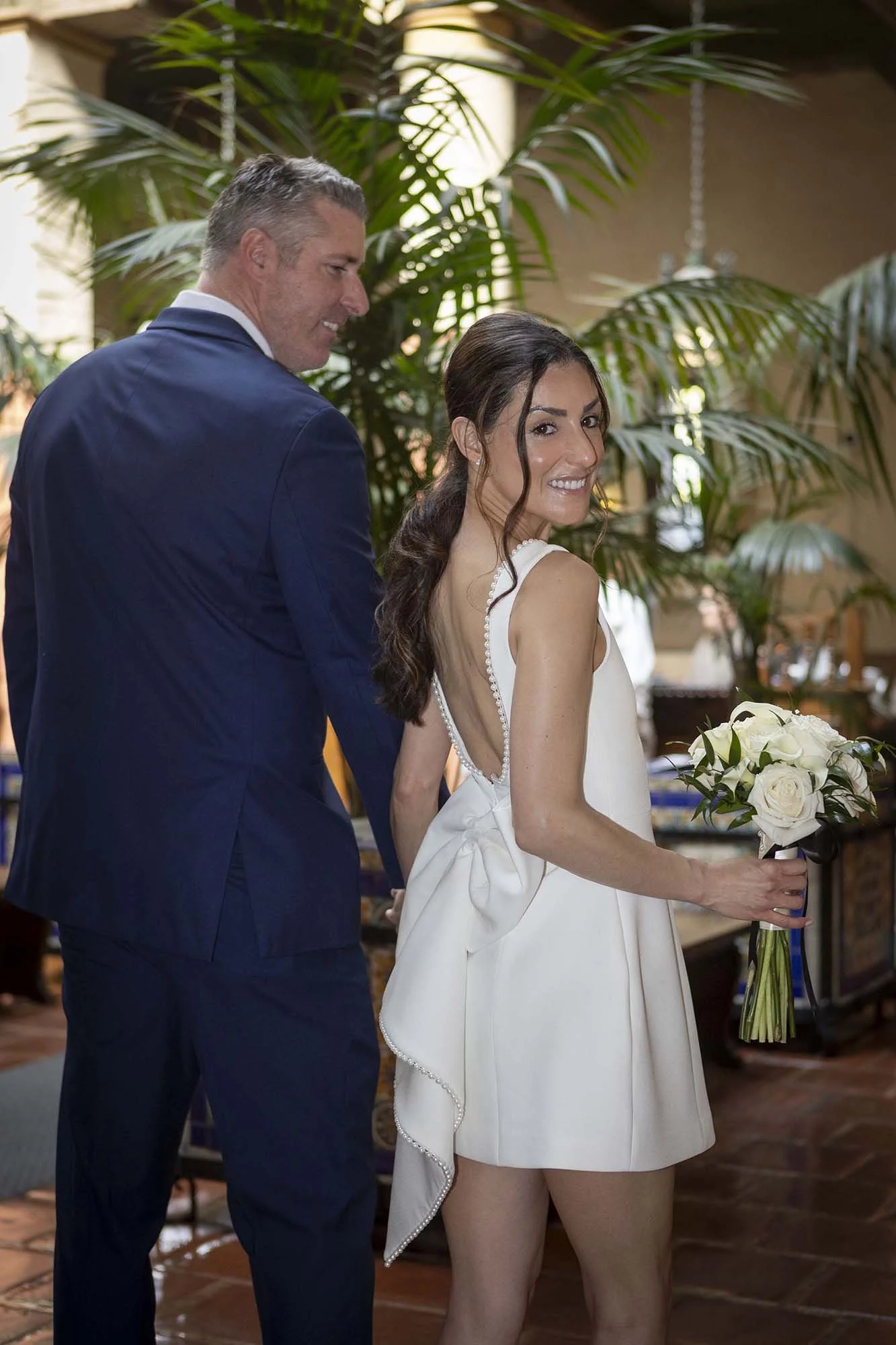 A bride holding a bouquet of white roses, wearing a white dress with a low back and bow details, smiling at the camera; a groom in a dark suit standing next to her, smiling; indoor setting with large green plants in the background.