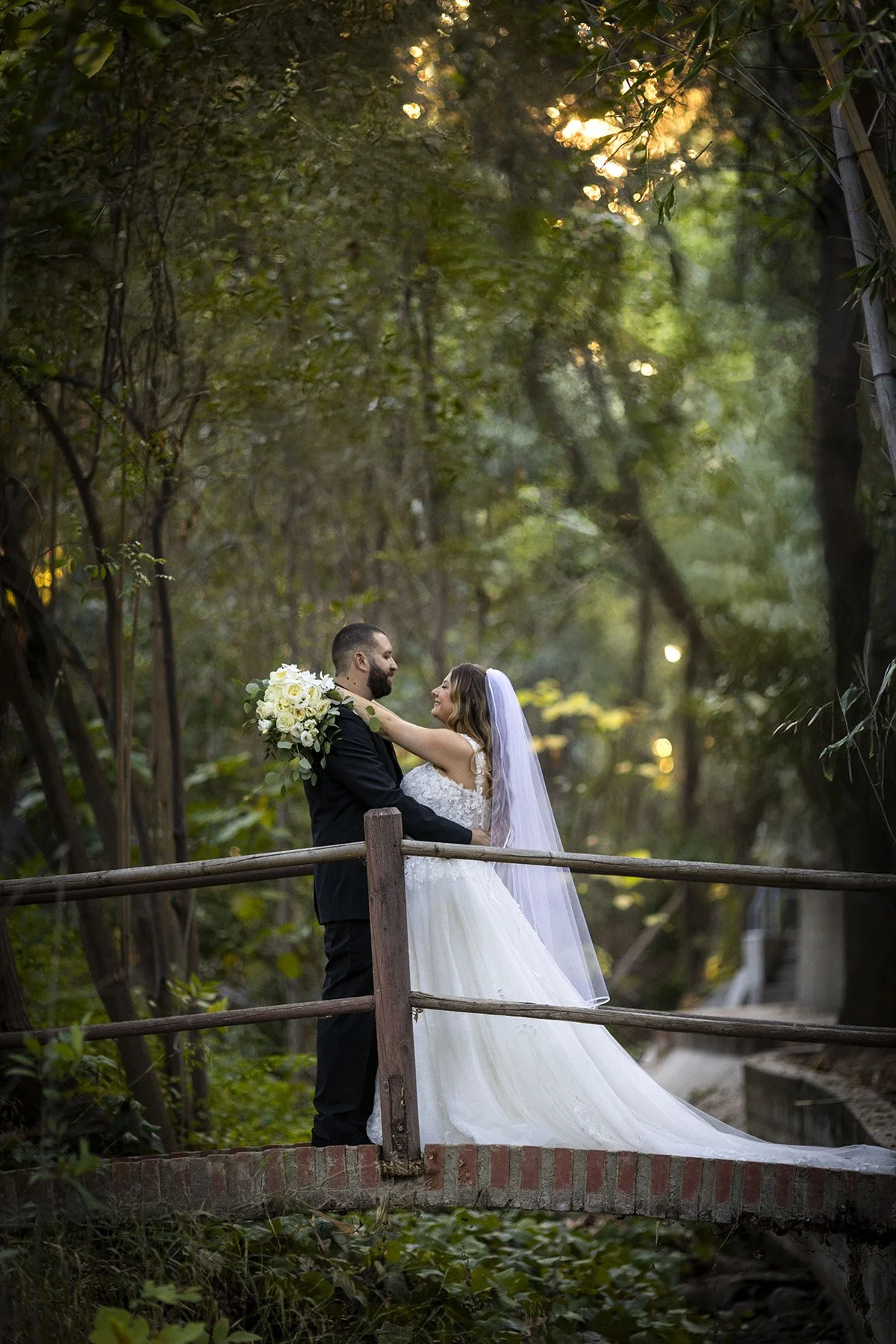A bride and groom sharing a romantic moment on a small wooden bridge in a wooded area during daylight. The bride is wearing a white wedding gown with a long veil and holding a bouquet, while the groom is dressed in a black suit. They are smiling at each other amidst lush green trees and dappled sunlight.