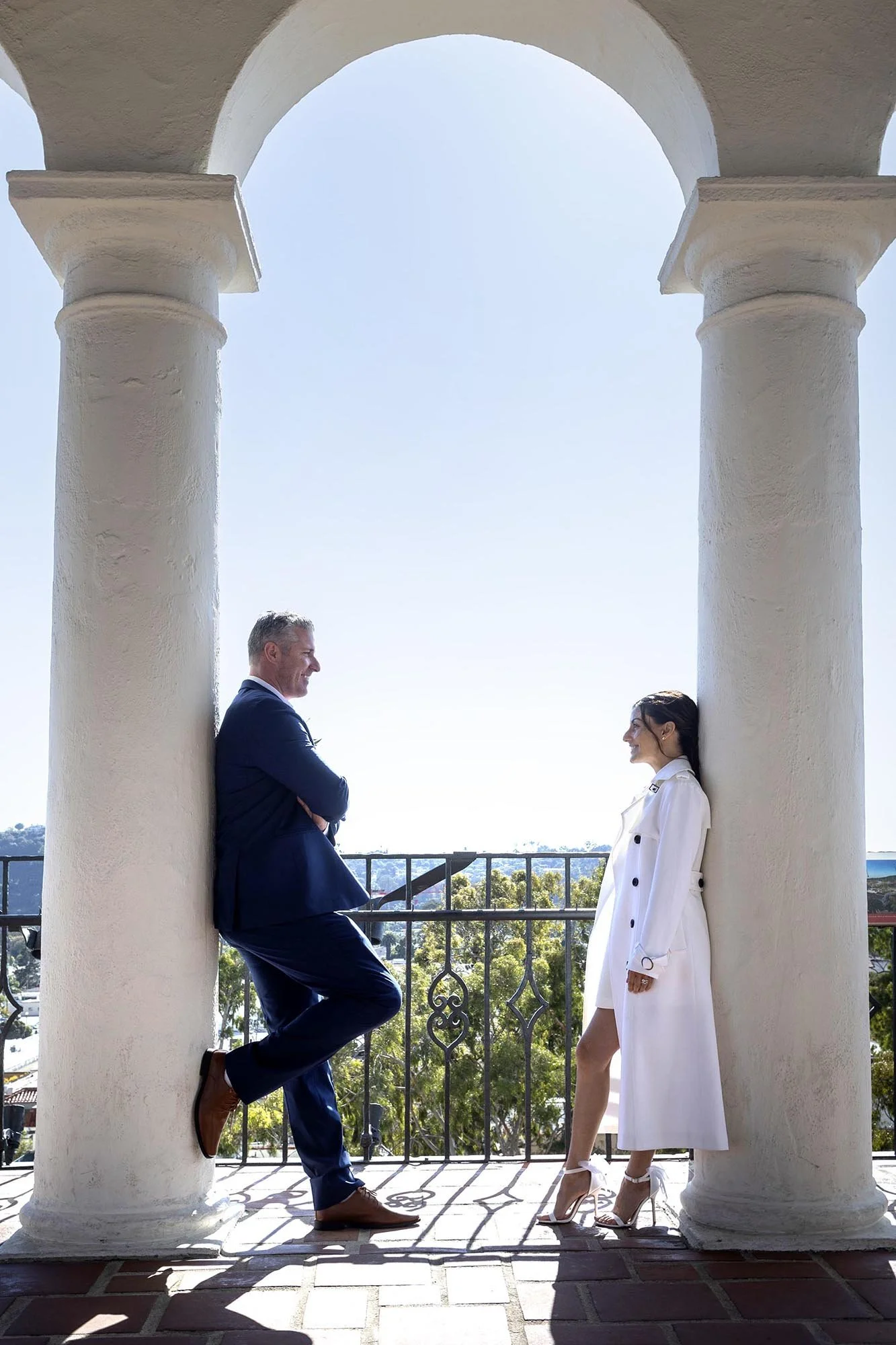 A man and woman in formal attire standing and leaning against large white columns on a balcony, smiling at each other with a scenic view of trees and clear sky in the background.